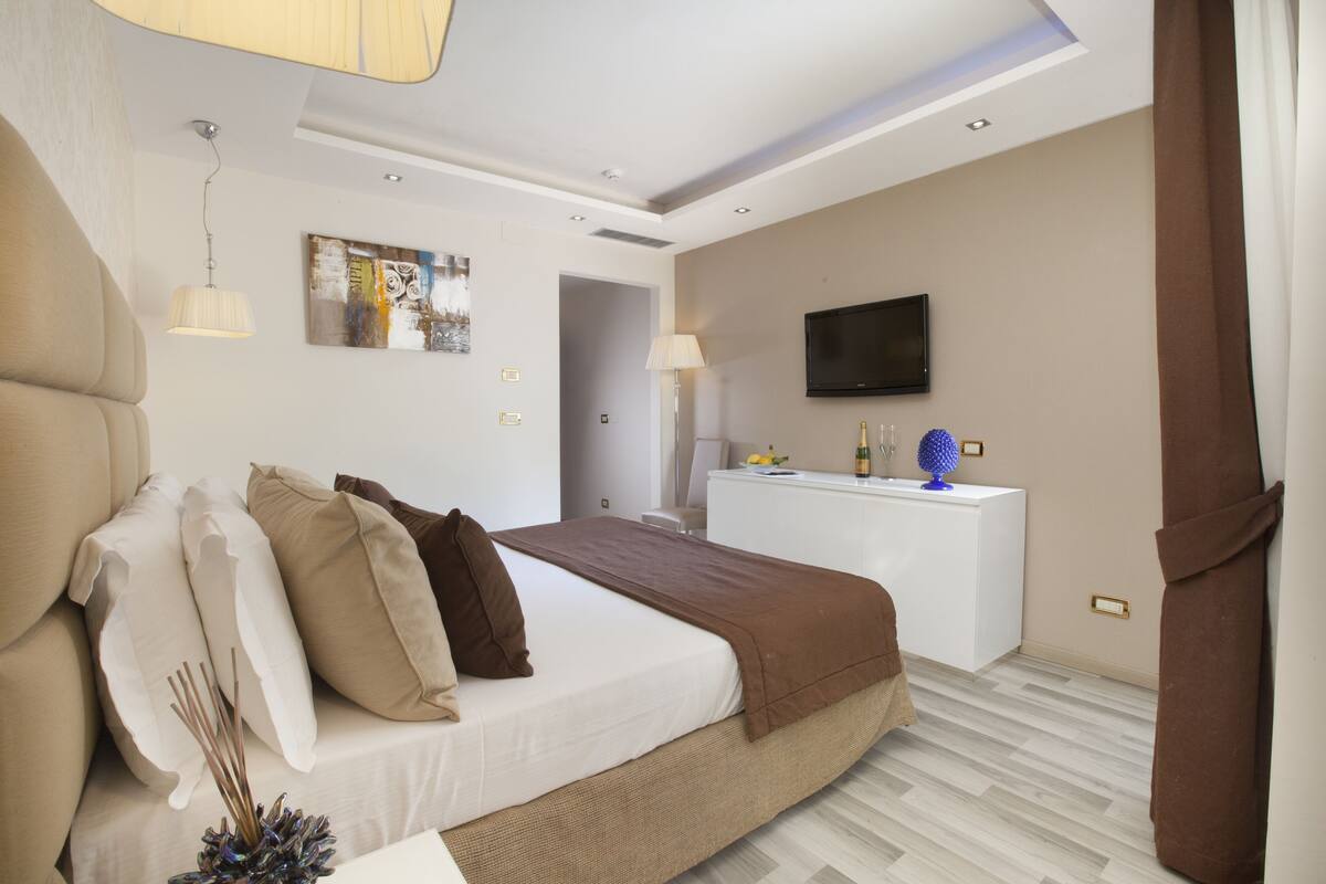 A modern hotel room with a large bed, a white desk, a flat-screen TV, and a painting on the wall.