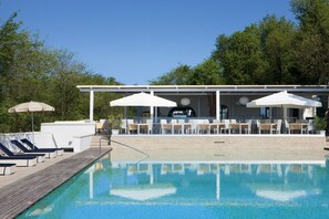 Zona de piscina amb gandules i ombrel·les, zona d'estar coberta amb taules, i vista a arbres al fons.