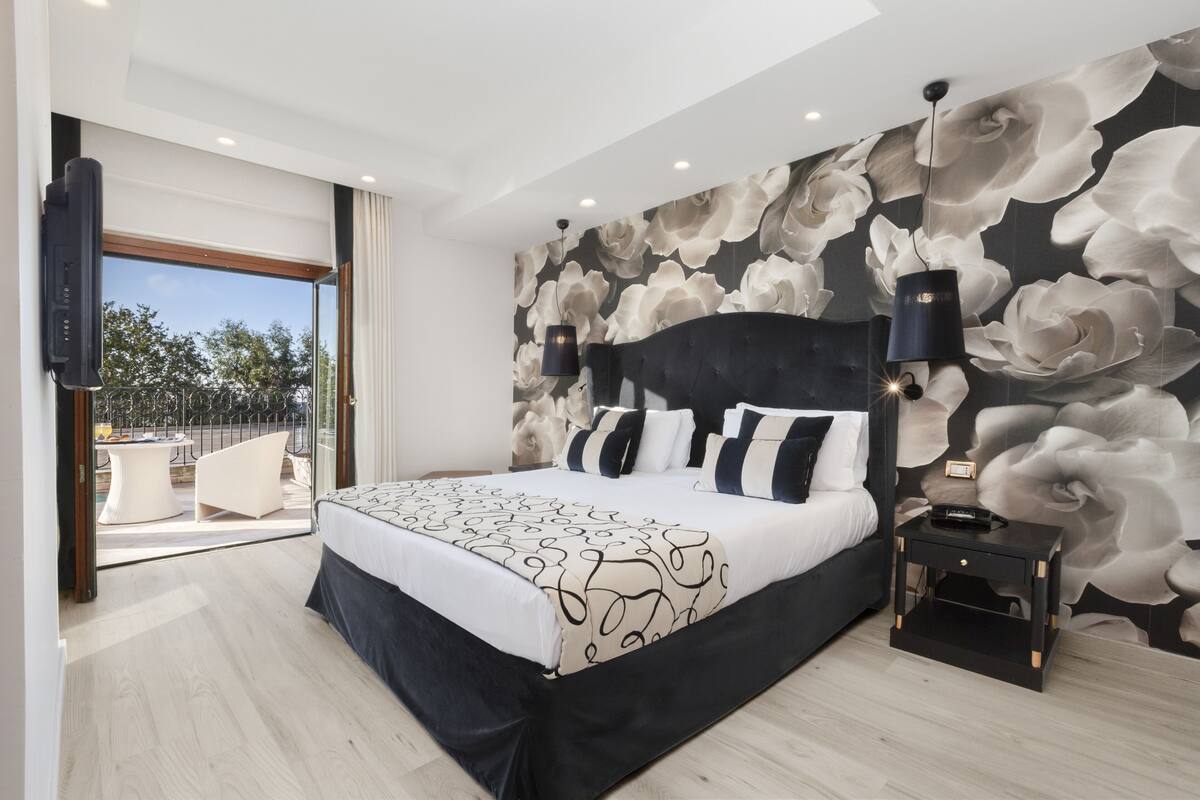 A modern bedroom with a large bed, a nightstand, a wall-mounted TV, and a balcony with outdoor furniture.