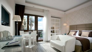 A modern hotel room with a bed, a desk with a computer, a chair, and a balcony with a view of palm trees.