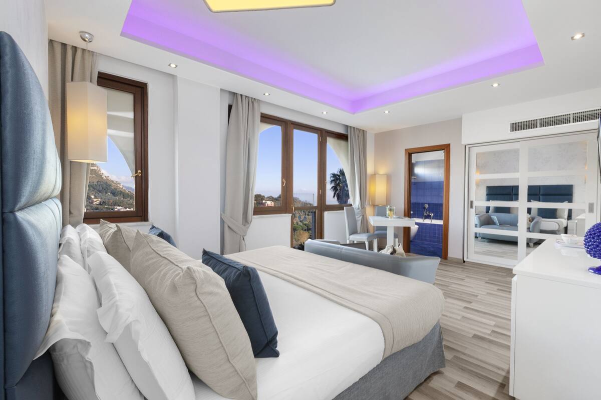 A modern hotel room with a large bed, a desk, and a view of the sea.