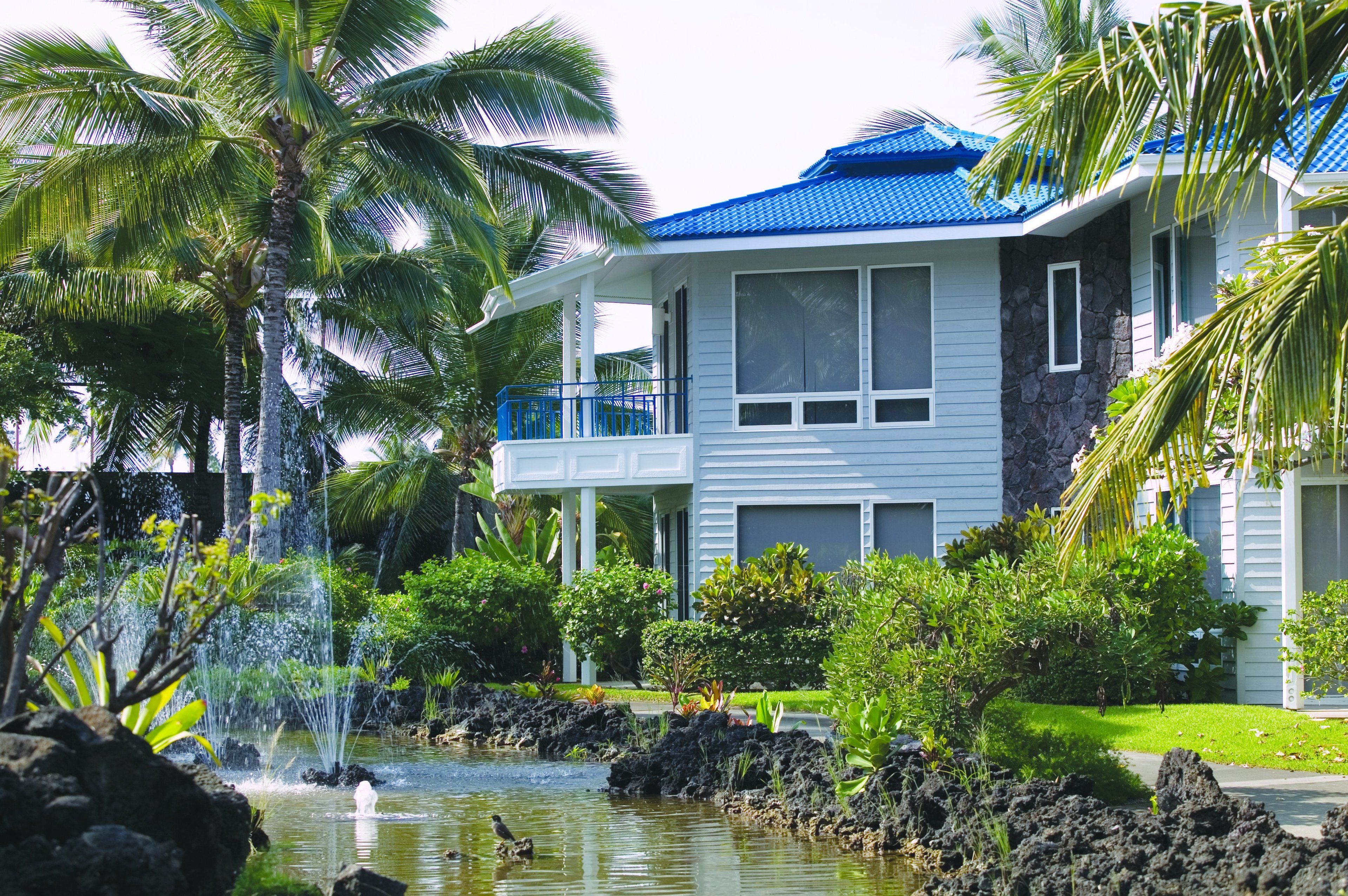 A two-story building with a blue roof, surrounded by lush greenery and a small pond with a waterfall.