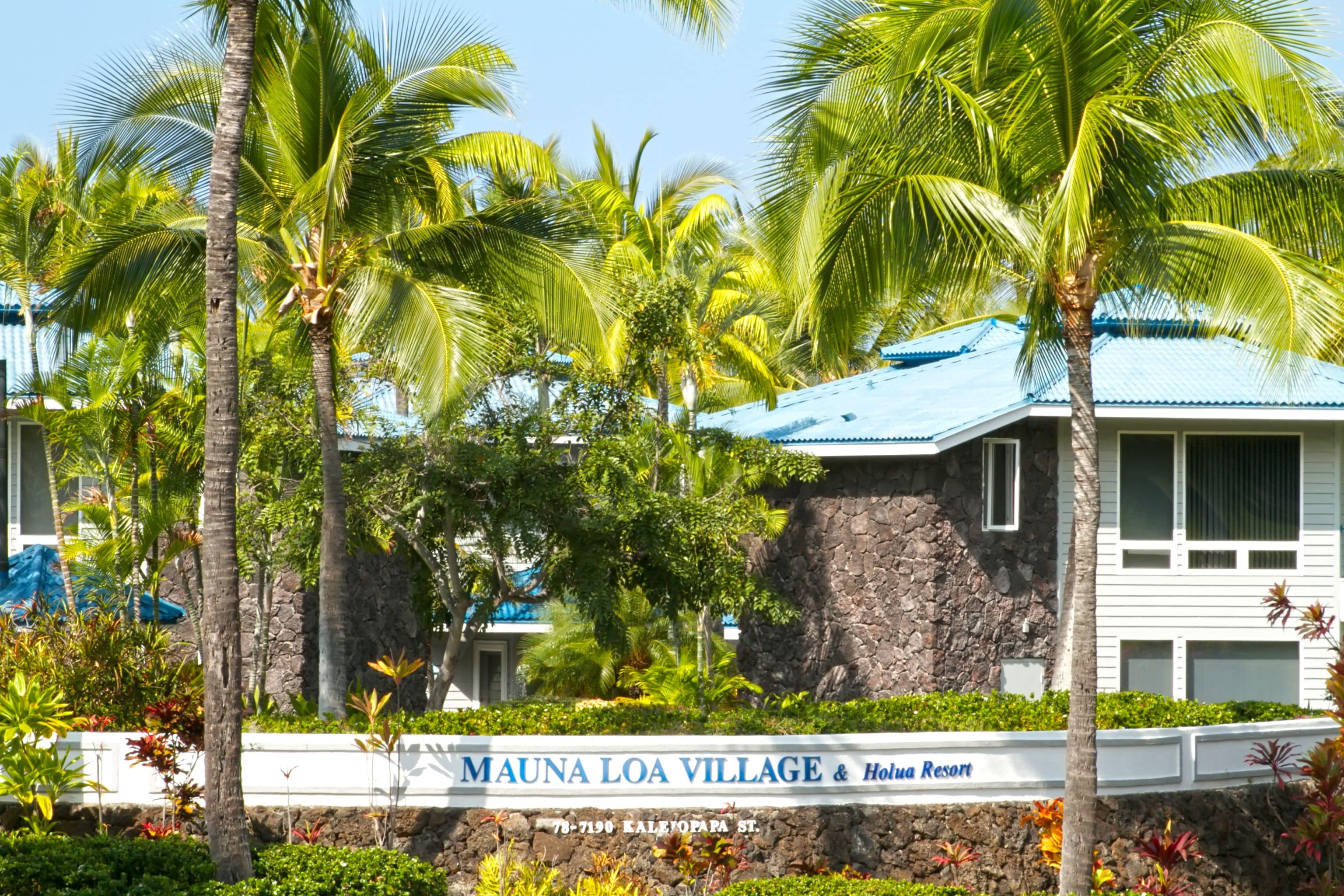 A sign for Mauna Loa Village & Holua Resort with a stone wall and palm trees.