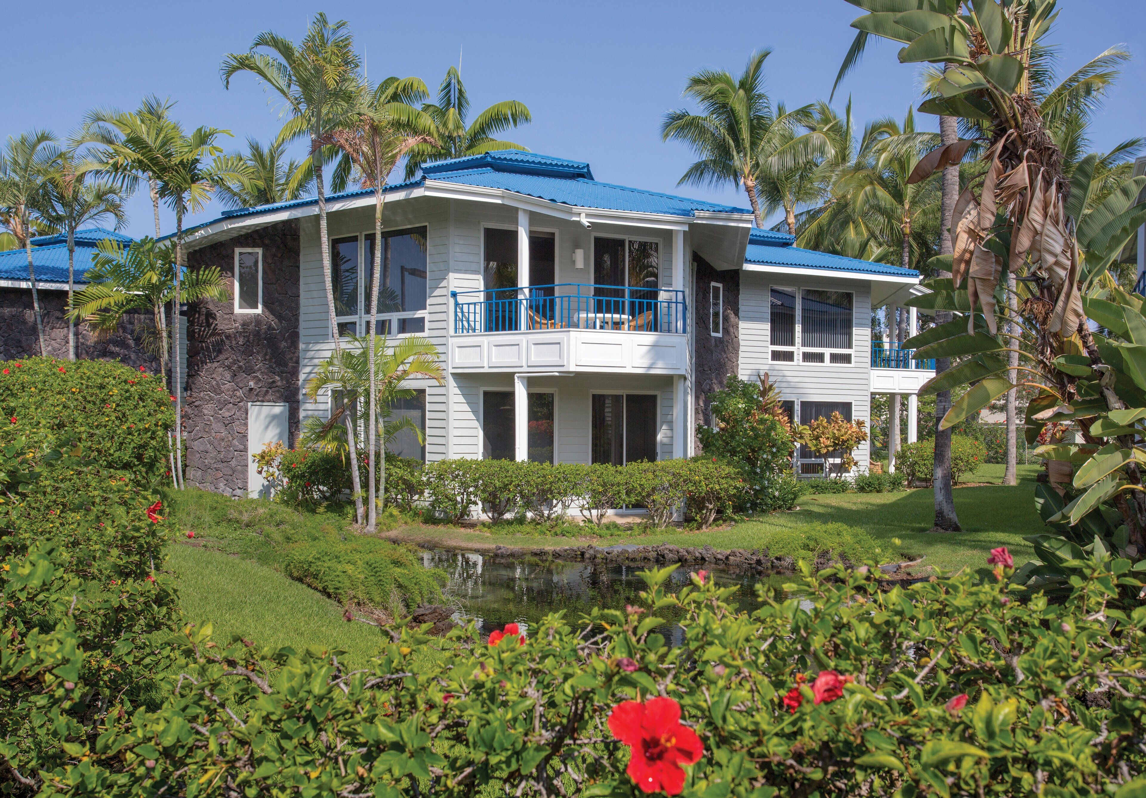A two-story house with a blue roof, surrounded by lush greenery and a small pond.