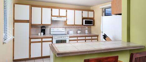 Cocina compacta con estufa, microondas y tostadora. Hay una ventana con persianas, un refrigerador y una zona de comedor con sillas.