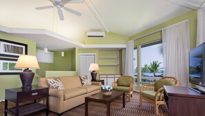 A hotel room with a sofa, two armchairs, a coffee table, a TV, a ceiling fan, an air conditioning unit, a window with curtains, and a patterned carpet.