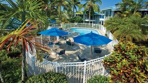 A pool area with lounge chairs and umbrellas, surrounded by palm trees and a white fence.