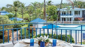A round table with four chairs, a vase with white flowers, and blue glasses on a balcony overlooking a pool and palm trees.
