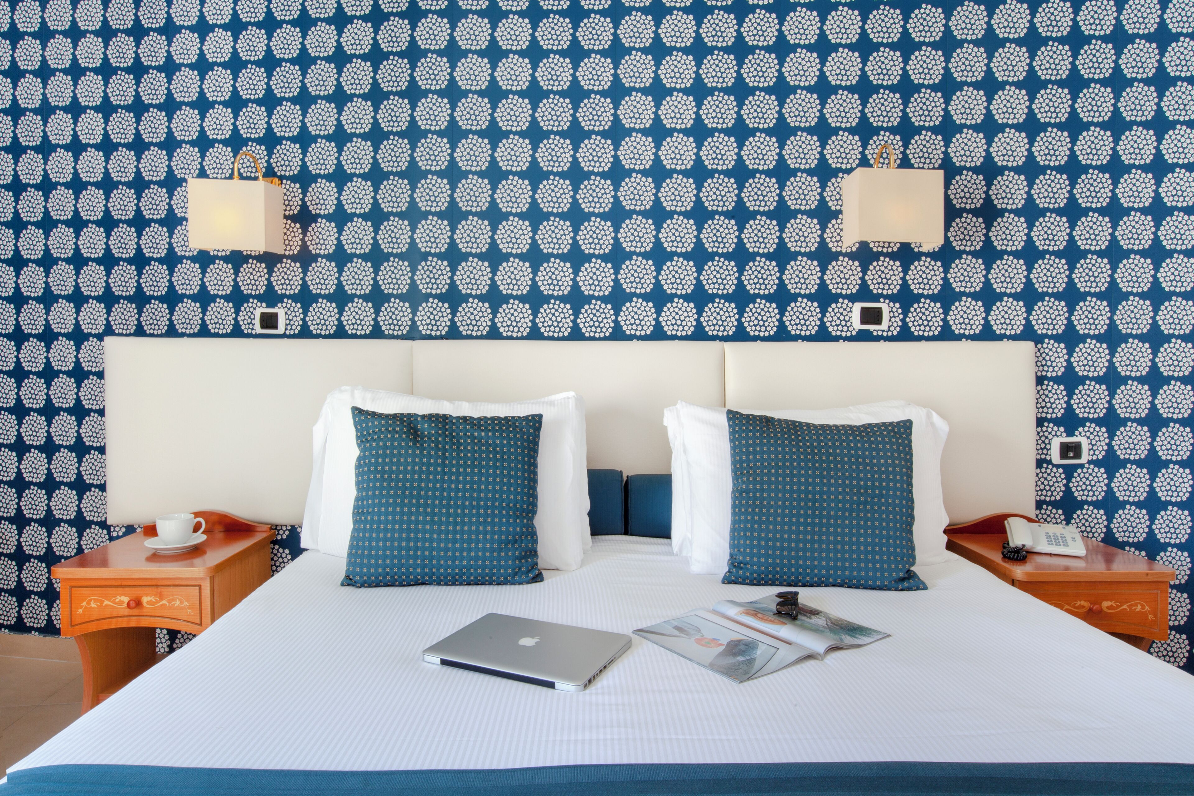 A hotel room with a bed, two pillows, a laptop, a magazine, a cup, and a phone on a nightstand.