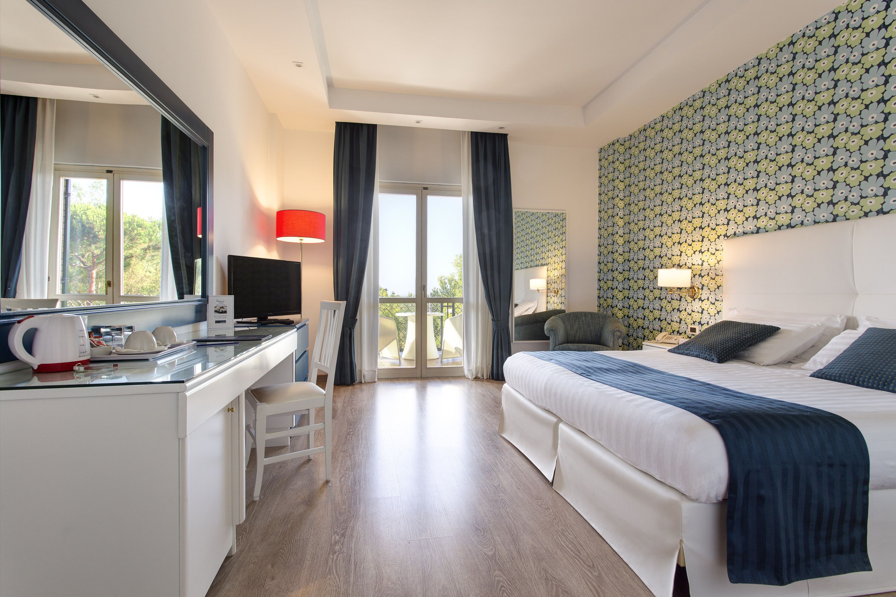 Une chambre d’hôtel comprenant un lit, un bureau avec un ordinateur, une chaise, une télévision et un balcon avec des rideaux.