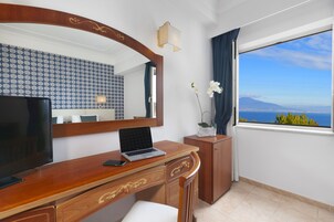 Camera d'albergo con una scrivania in legno, un computer portatile, una televisione, uno specchio, una finestra vista mare e un'orchidea in vaso.