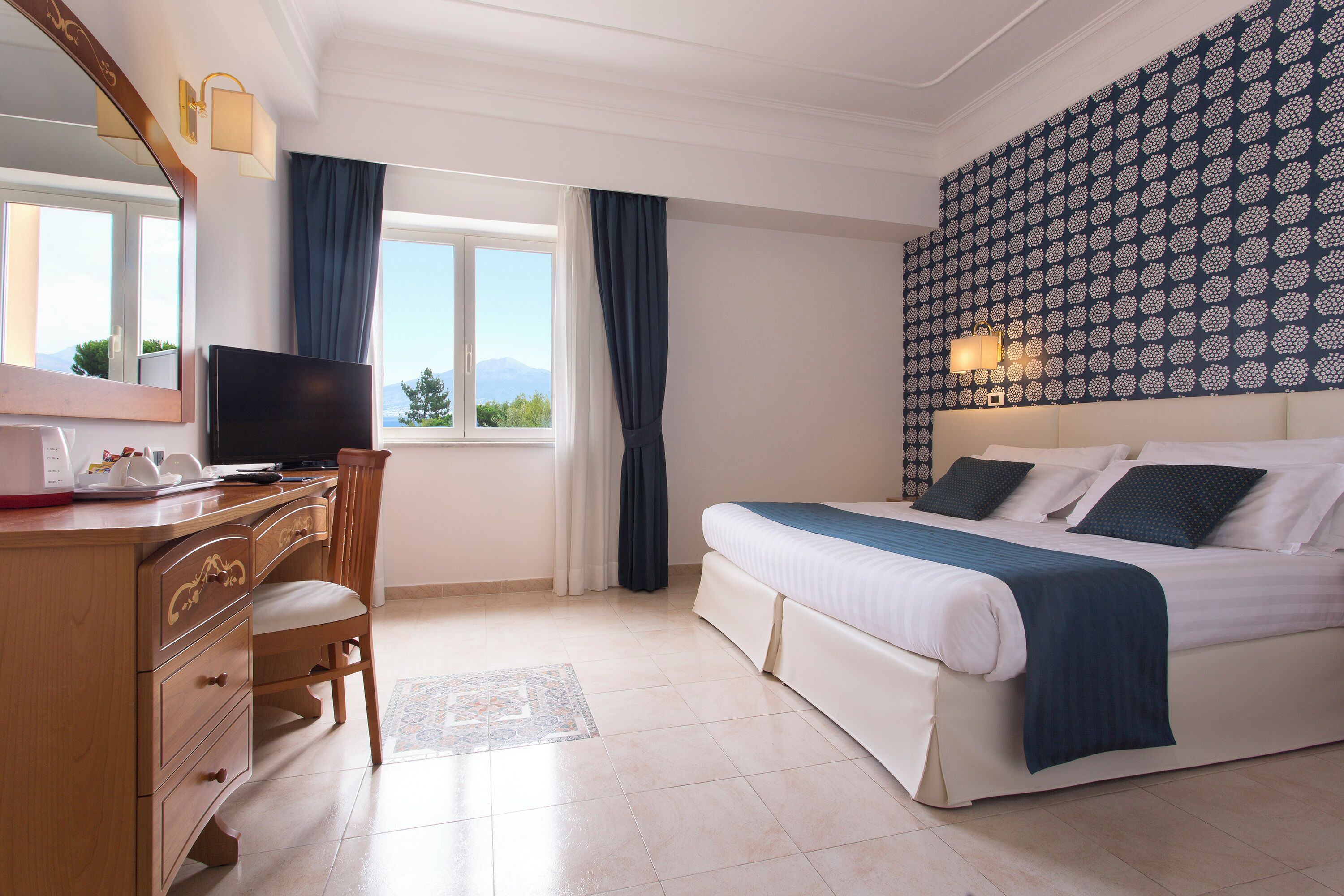 Camera d'albergo con un letto, una scrivania con televisore, una cassettiera e una finestra con tende.