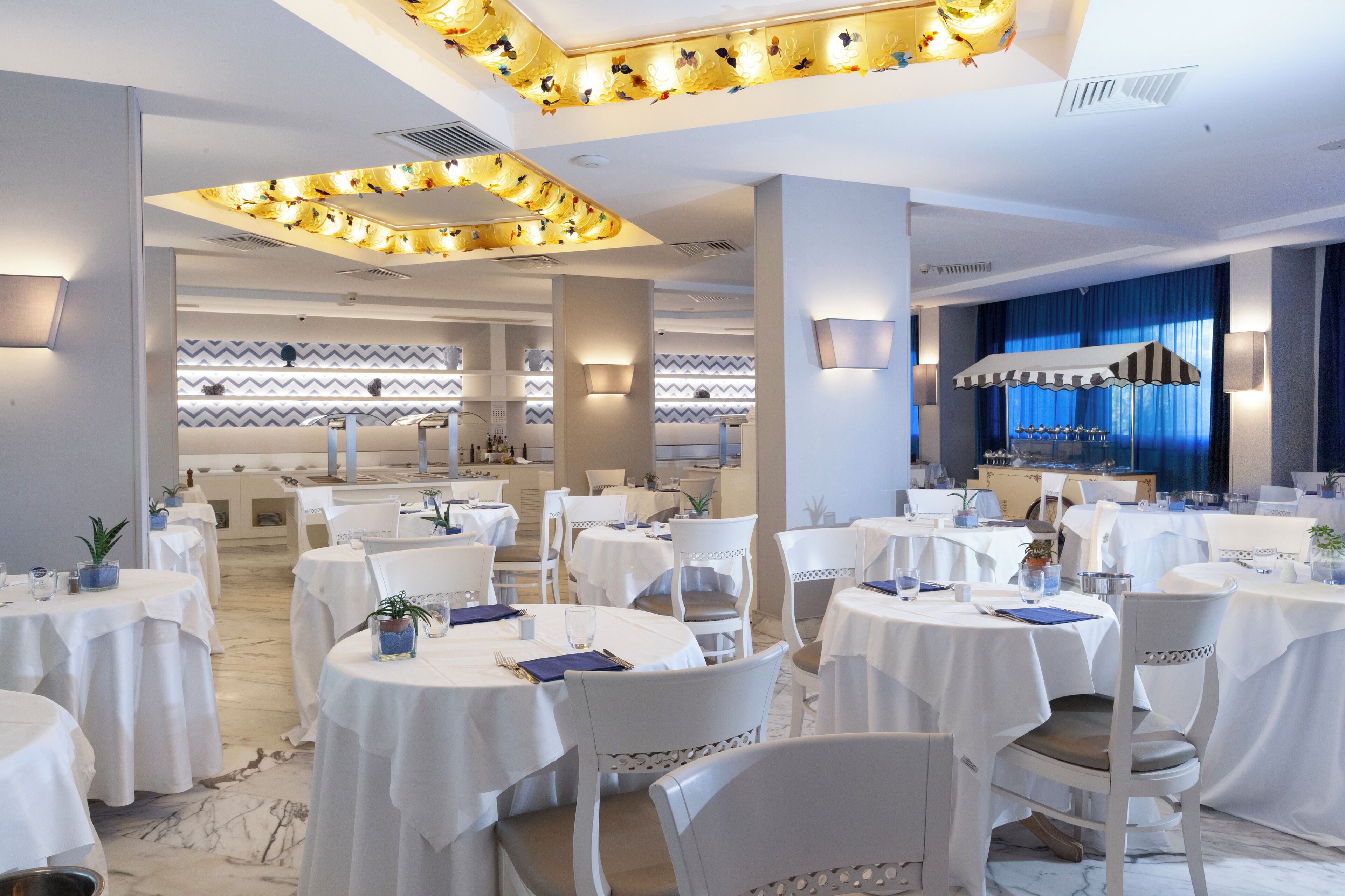 A hotel dining area with round tables, white chairs, and a central bar area with blue curtains.