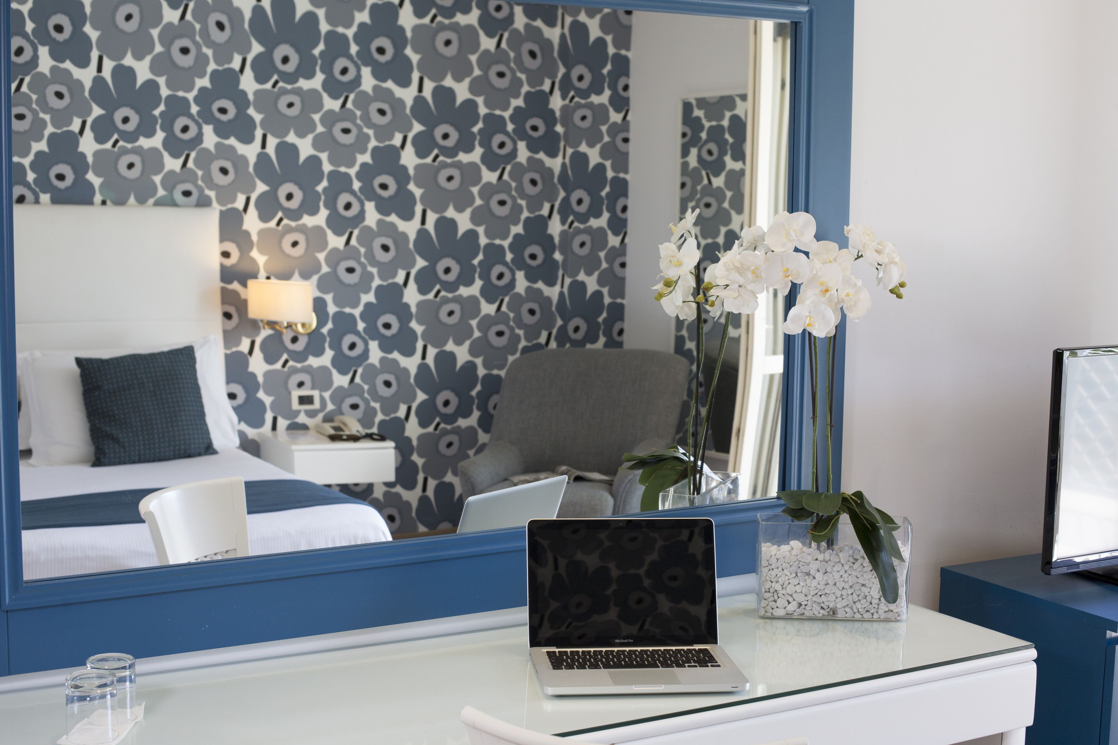 Een hotelkamer met een bed, een bureau met een laptop, een stoel, een vaas met witte bloemen en een grote spiegel die de kamer weerspiegelt.