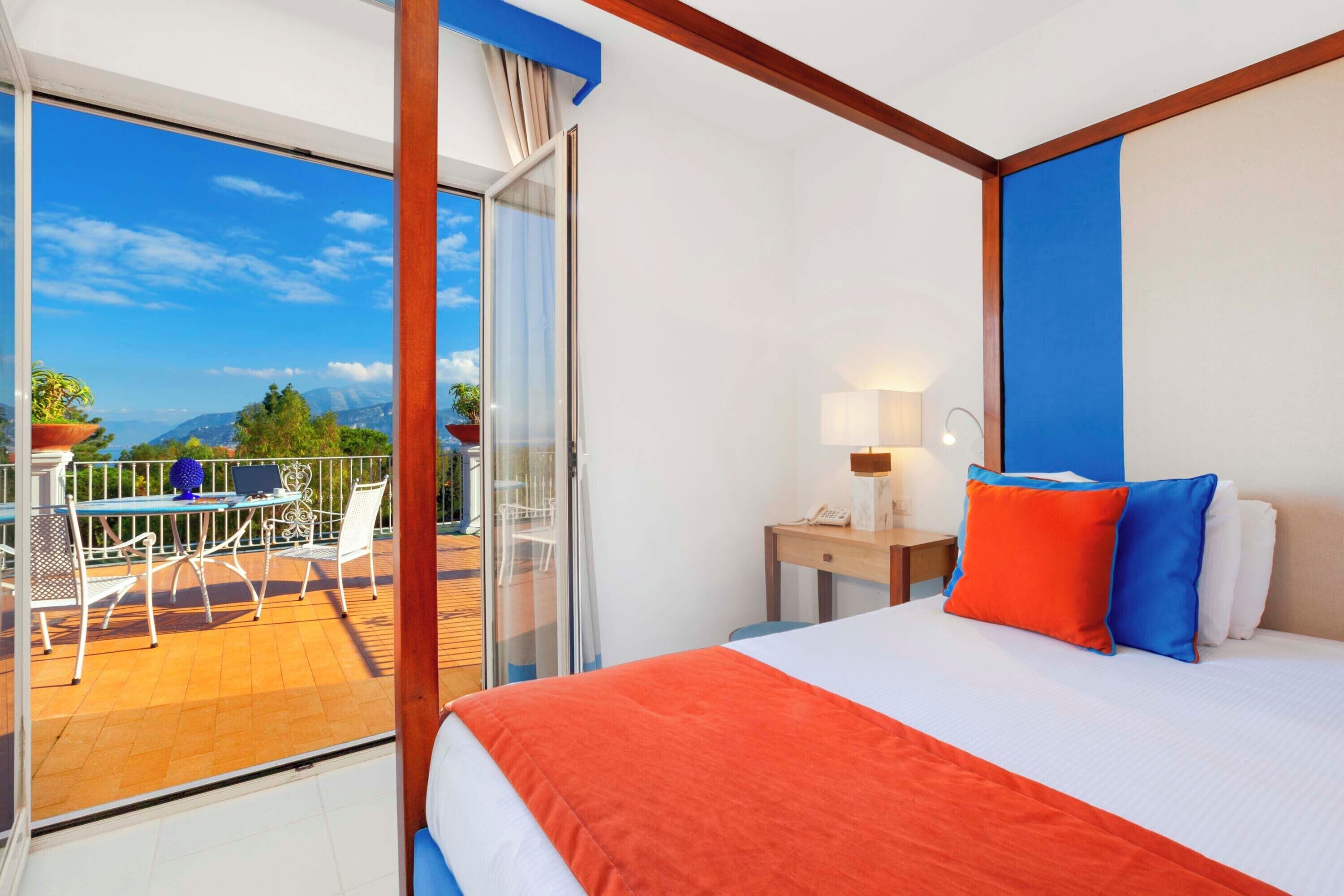 Une chambre d’hôtel avec un grand lit, un balcon doté d’une table et de chaises, et une vue sur les montagnes.