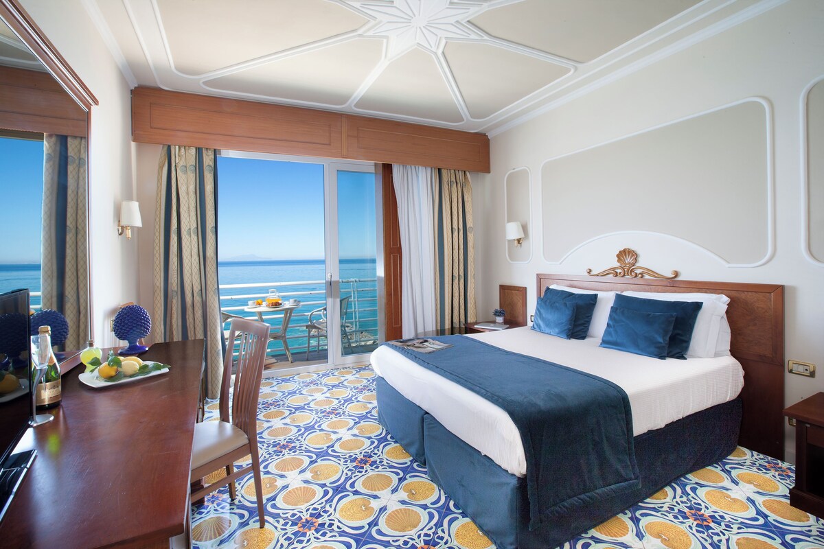 Quarto de hotel com uma cama grande, uma mesa com cadeira e vista para o mar.
