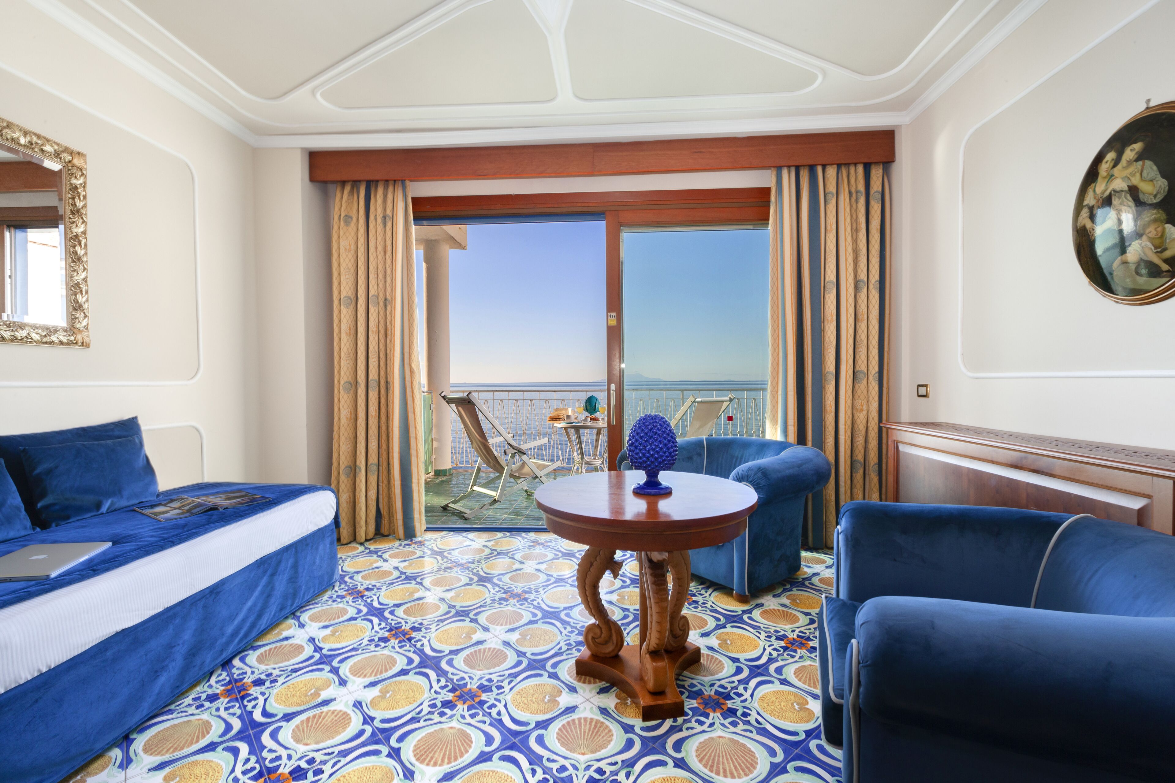 A hotel room with a bed, two blue armchairs, a small round table, and a balcony with a view of the sea.