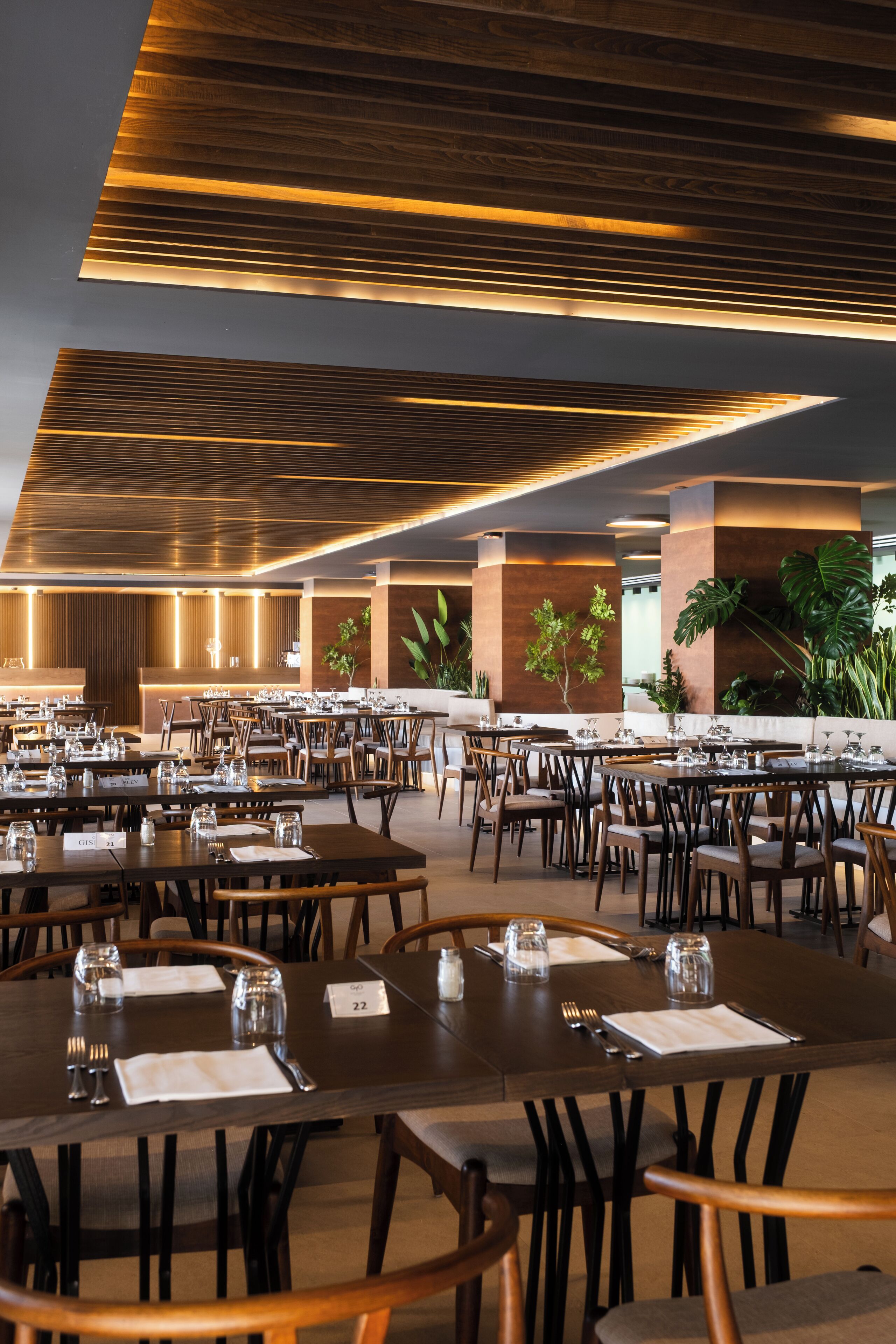 A spacious restaurant with multiple dining tables and chairs, a modern ceiling design, and indoor plants.