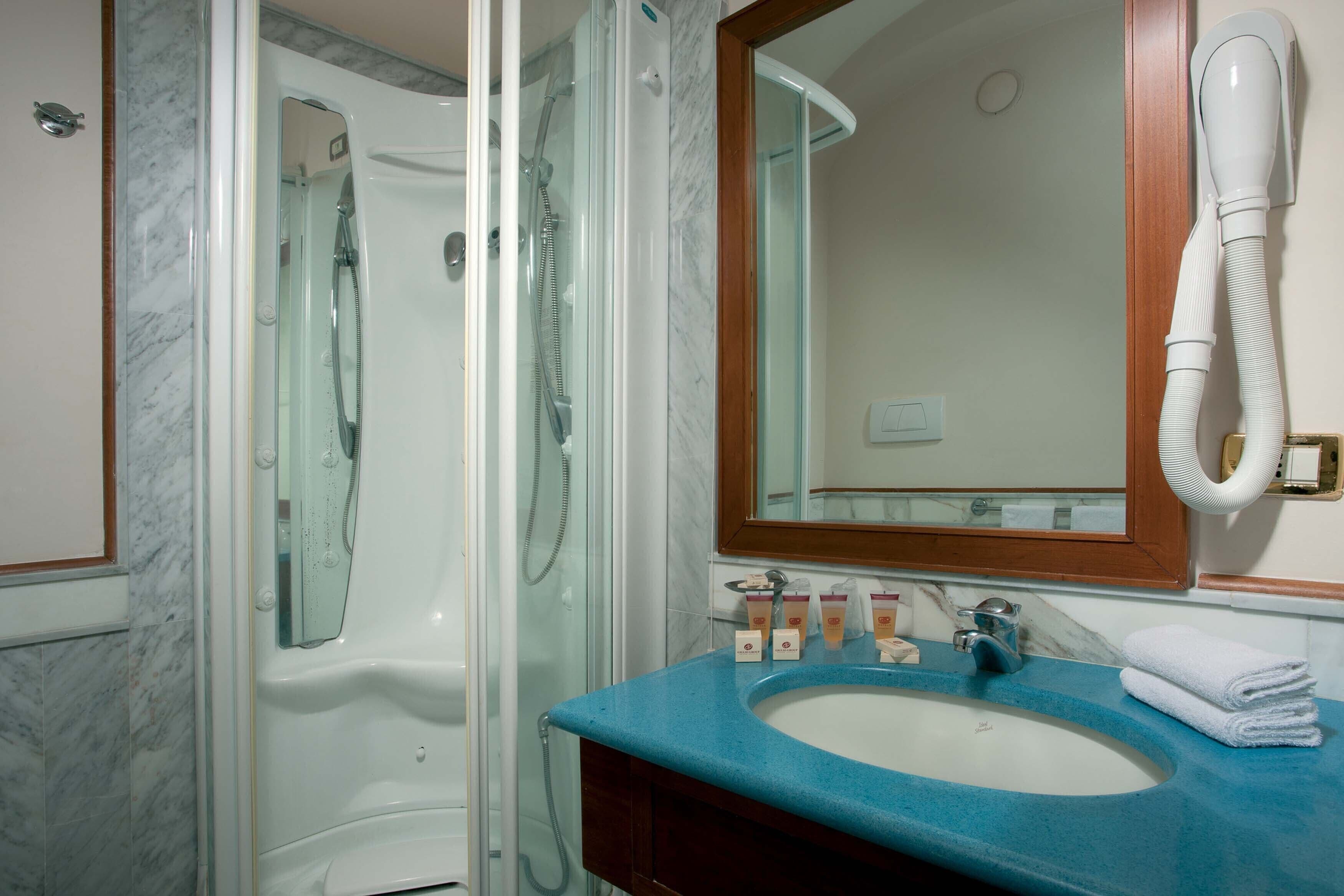 A bathroom with a glass-enclosed shower, a blue sink, and a mirror with a hairdryer mounted on the wall.