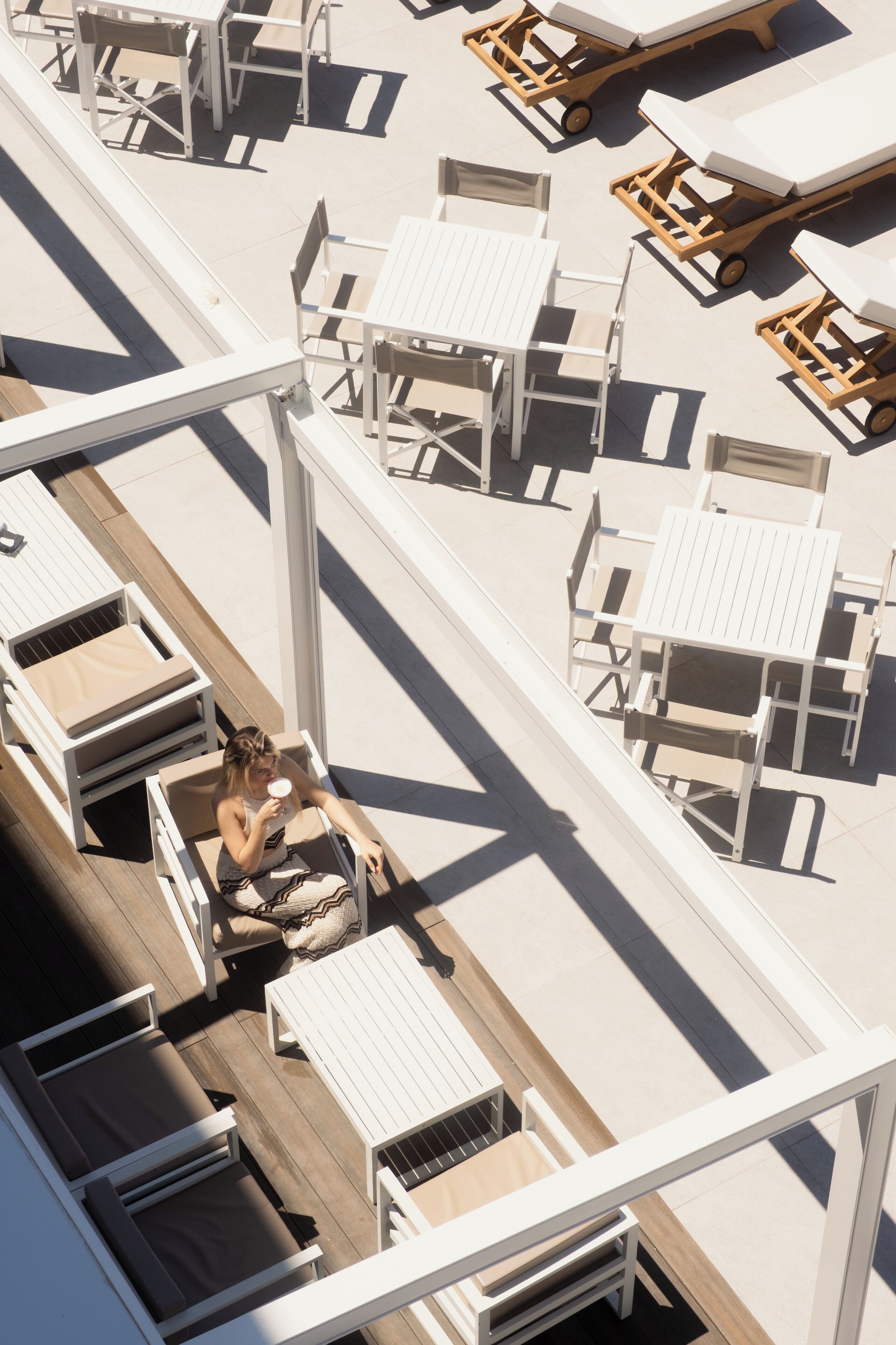 An aerial view of a rooftop lounge area with multiple seating arrangements, including sun loungers and tables, and a person seated at a table.