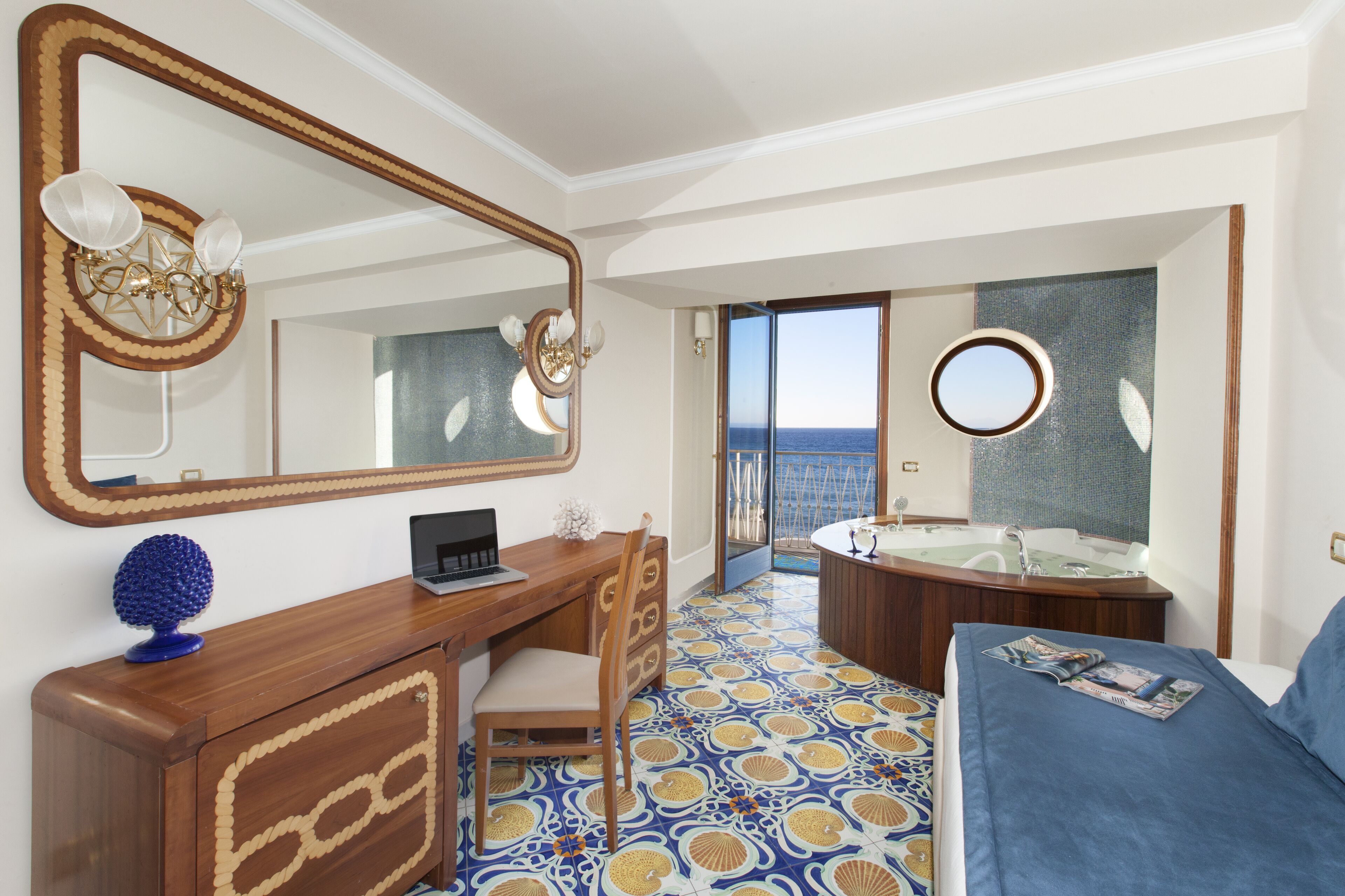 A hotel room with a wooden desk, a chair, a bed with a blue bedspread, a large mirror, a round mirror, a bathtub, and a view of the sea through a glass door.