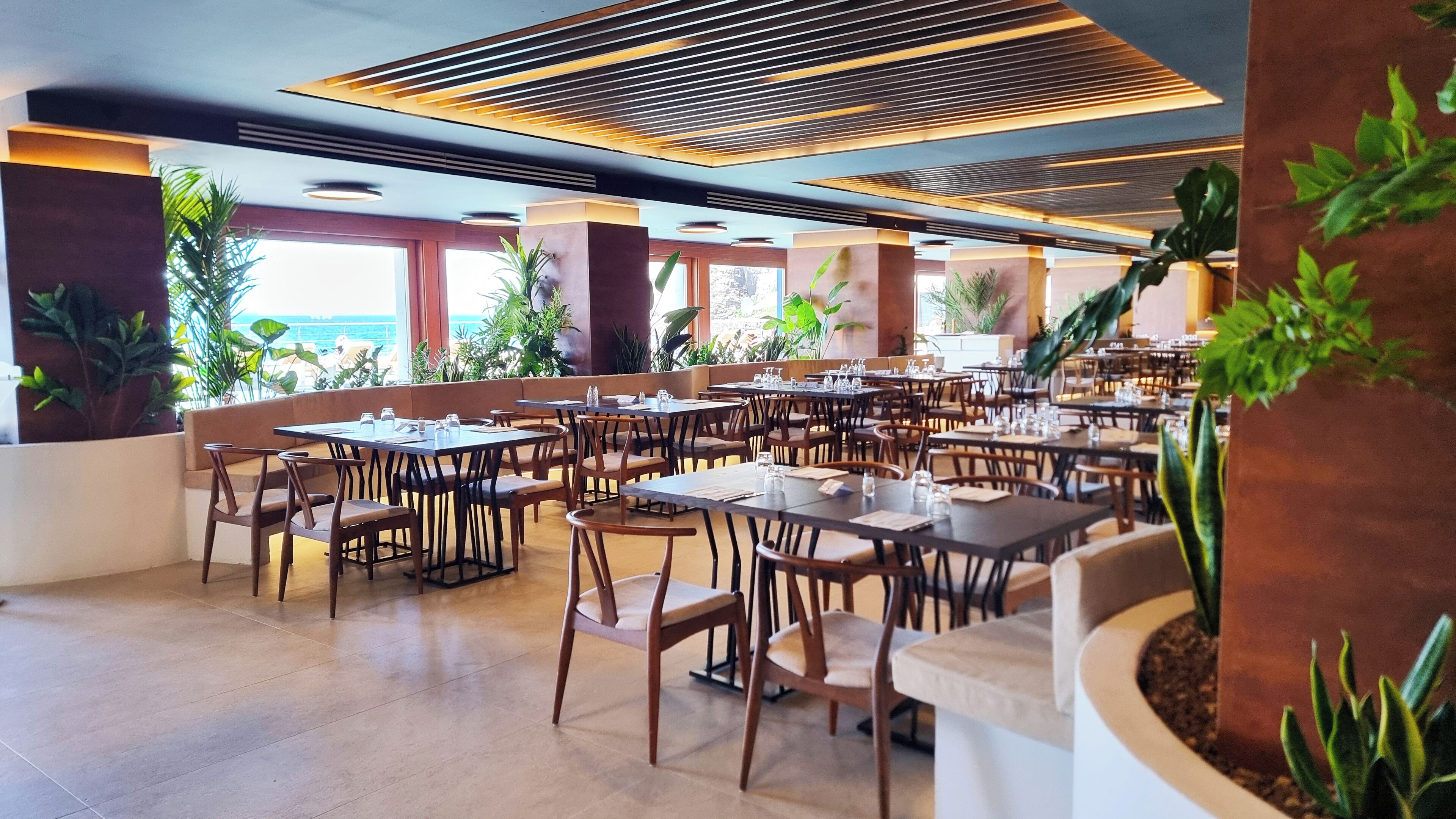 Um restaurante espaçoso com várias mesas e cadeiras, amplas janelas com vista para o mar e plantas dentro.