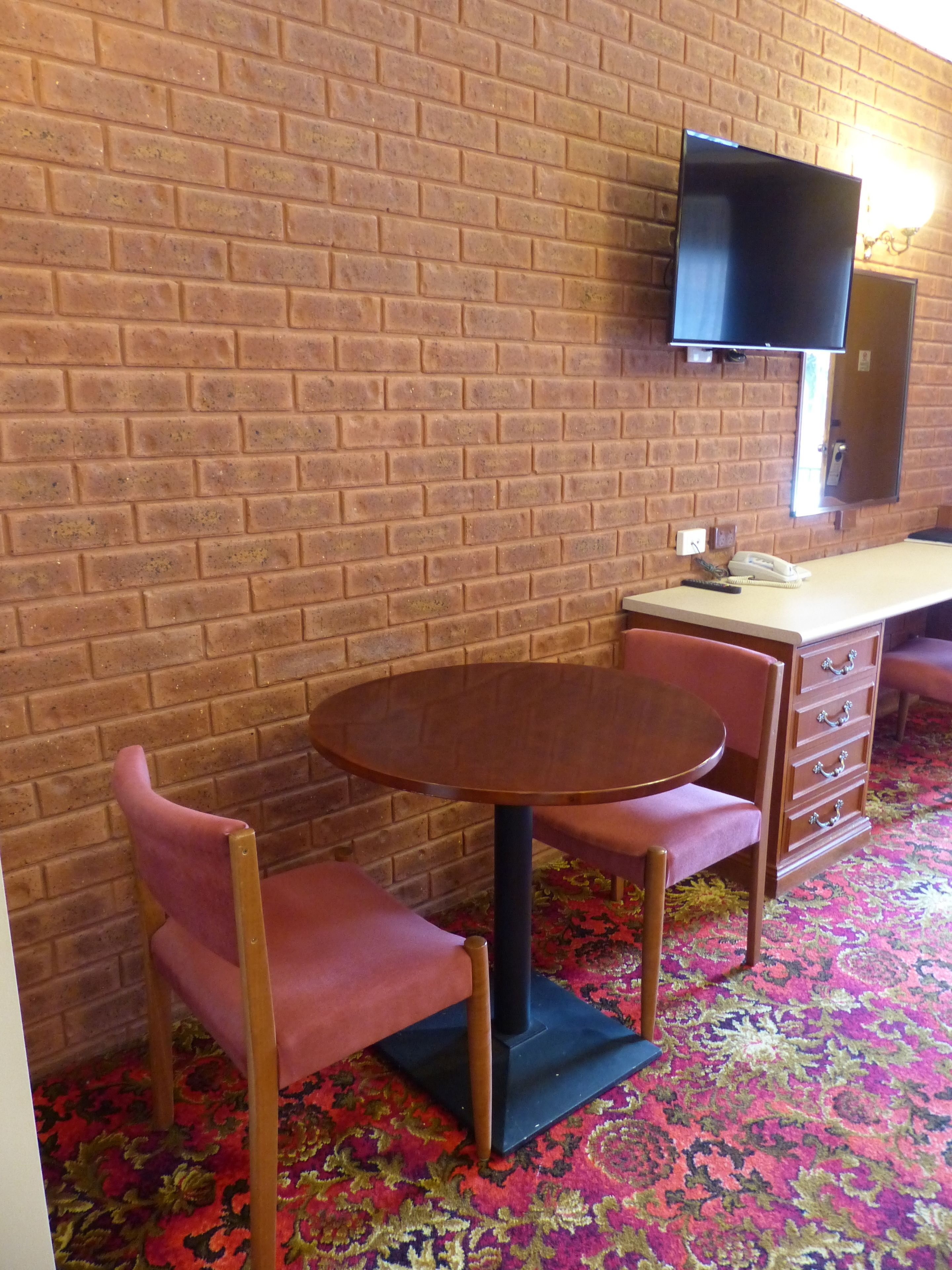 Una habitación con pared de ladrillo, una mesa redonda de madera con dos sillas, un televisor de pantalla plana montado en la pared y un teléfono sobre la mesa.