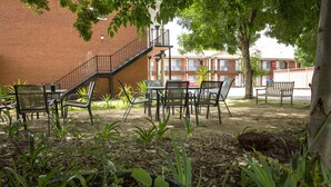 An outdoor patio area with tables and chairs, surrounded by plants and a brick building in the background. - Meramie Motor Inn (Albury)