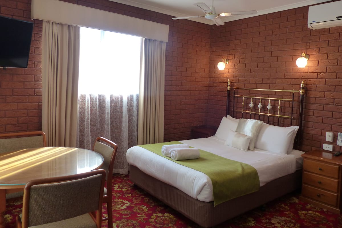 A hotel room with a brick wall, a bed with white linens, a green throw, a desk with a chair, and a television mounted on the wall.