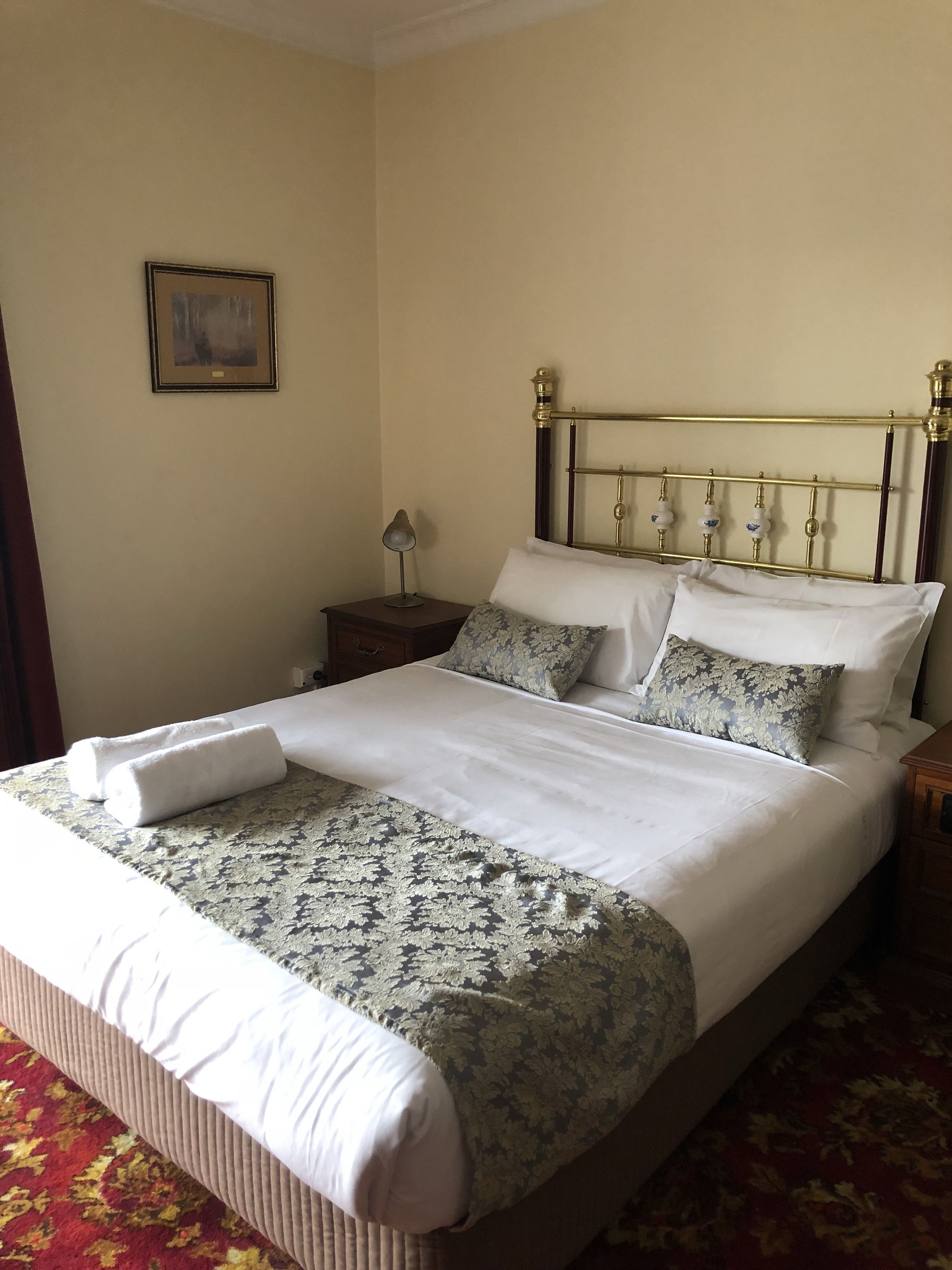 Una cama bien hecha con un edredón floral y dos almohadas, una lámpara de noche y un cuadro en la pared.