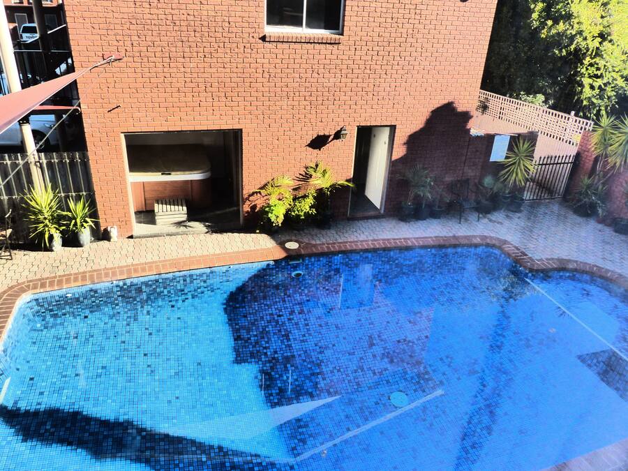 Une piscine dont le bord est revêtu de carreaux, entourée d’un bâtiment en briques doté de fenêtres et d’un petit espace extérieur agrémenté de plantes en pot.