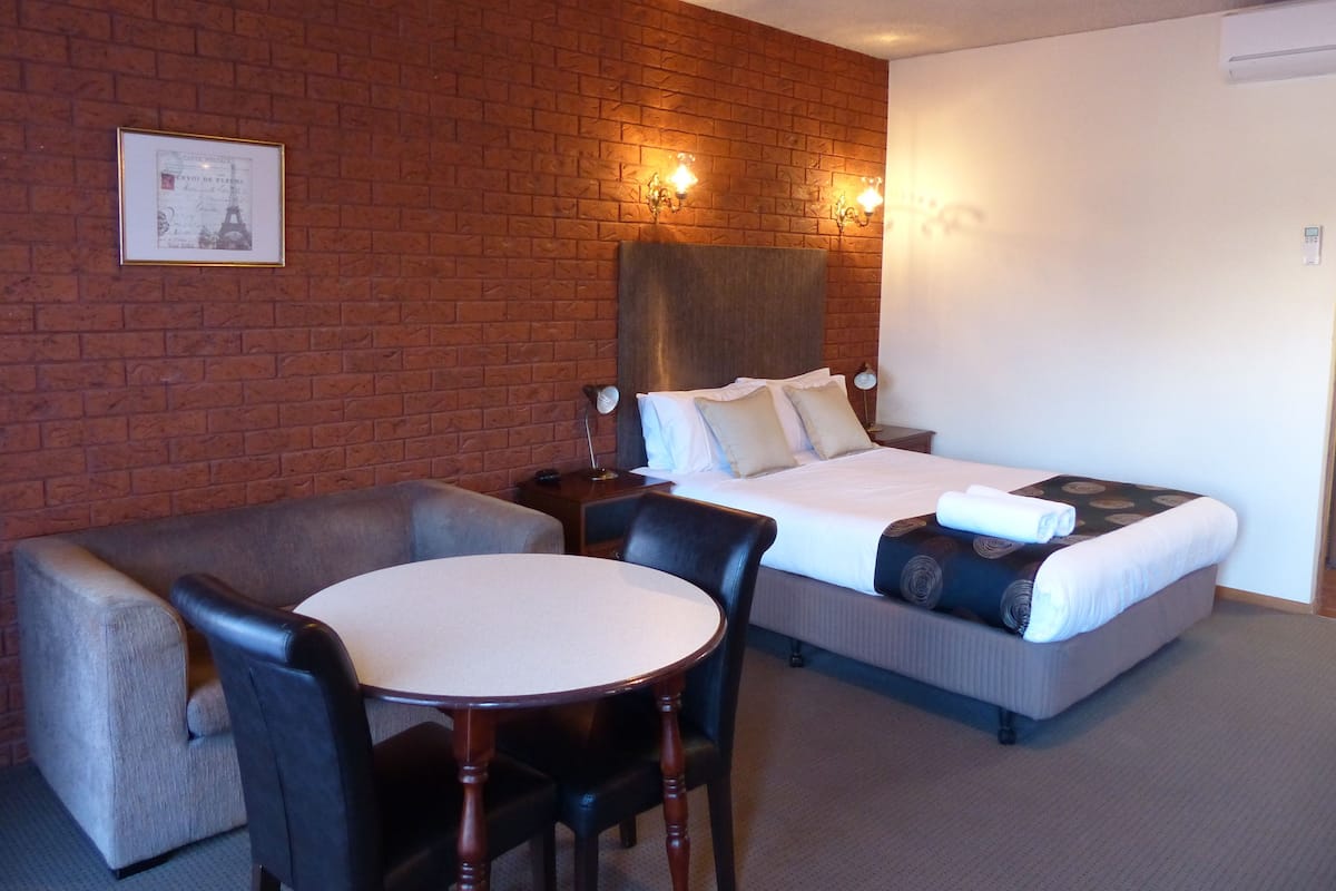 A hotel room with a brick wall, a bed with white linens, a round wooden table, and two armchairs.