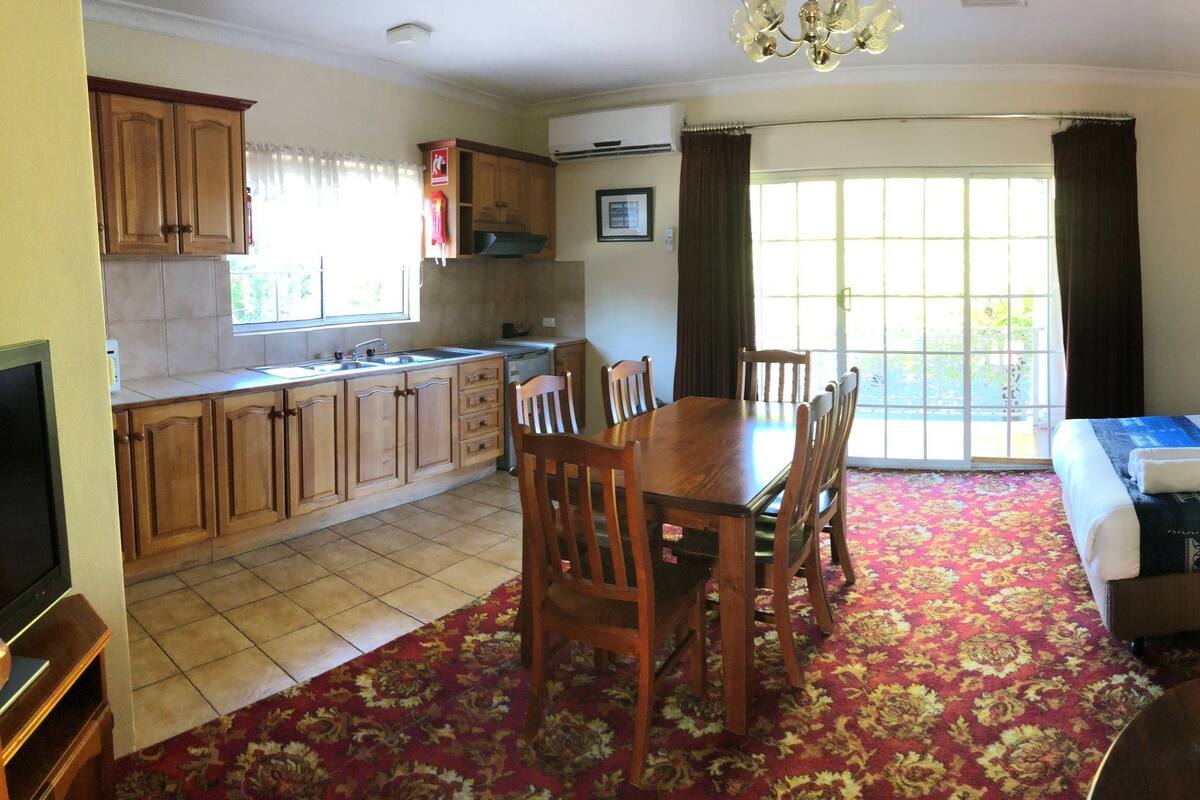 A compact hotel room with a bed, a sofa, a dining table with chairs, a kitchen area, and a large window.