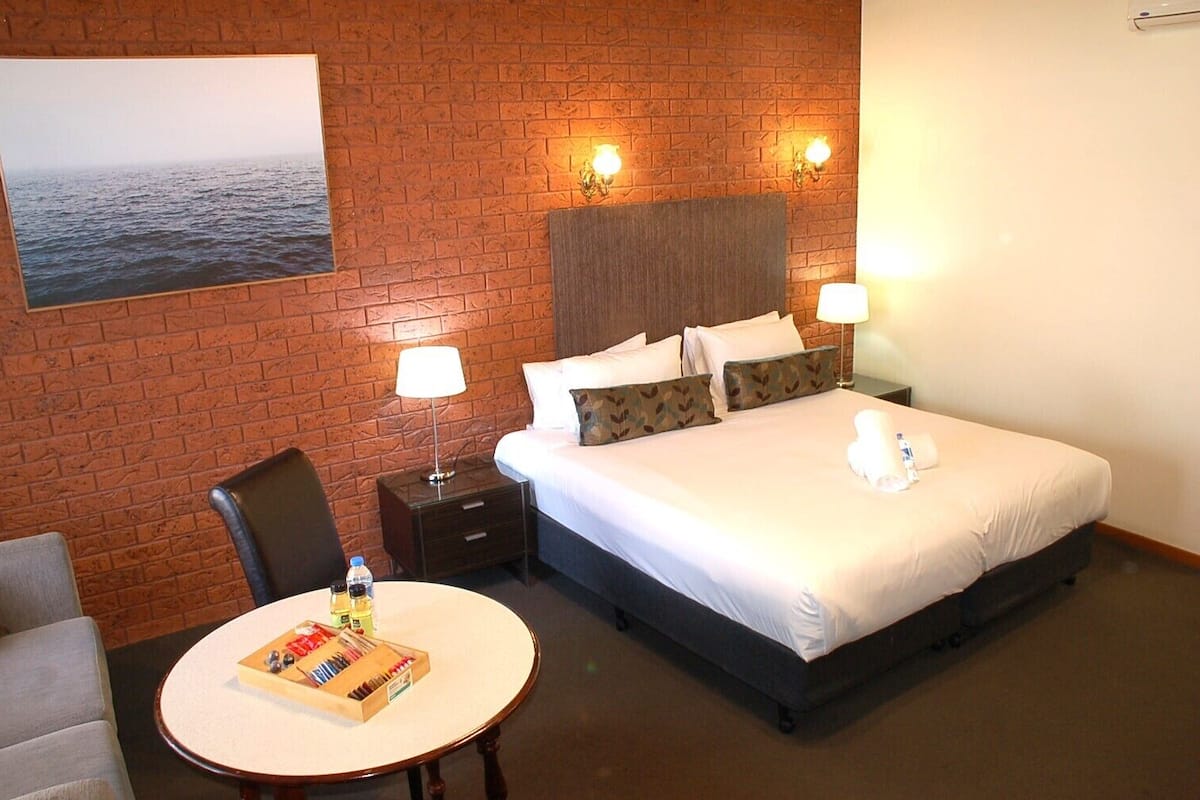 A hotel room with a brick wall, a bed with white linens, two bedside lamps, a nightstand, a table with a board game, and a chair.