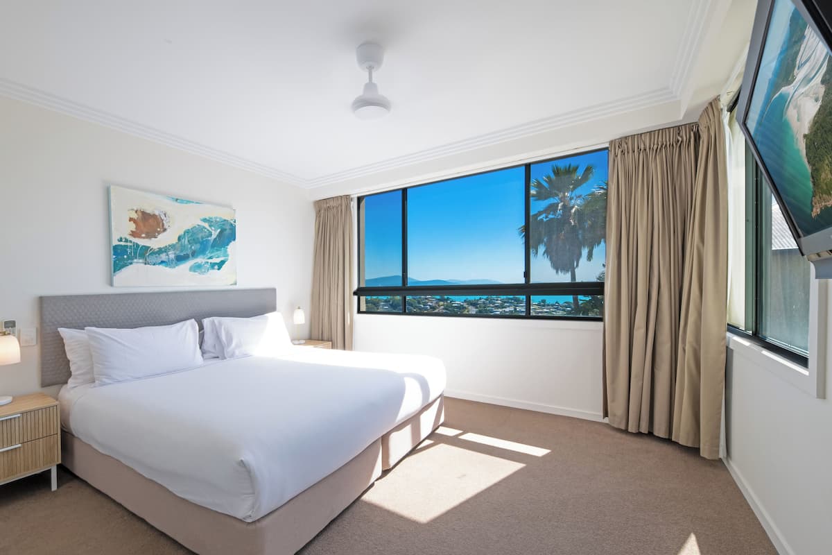 Habitación de hotel con una cama grande, una mesita de noche, un ventilador de techo, una ventana con vista a la ciudad y un cuadro en la pared.