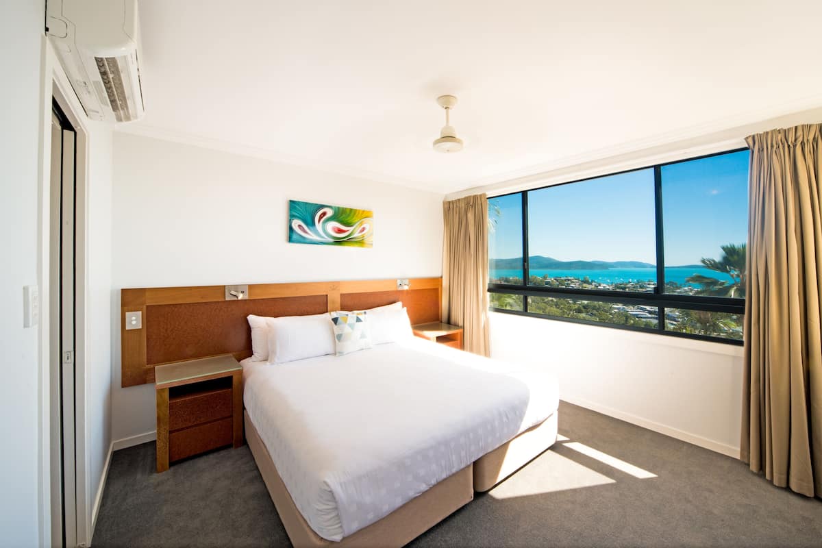 Habitación de hotel con una cama grande, una mesita de noche, un ventilador de techo, una ventana con vista al mar y un cuadro en la pared.