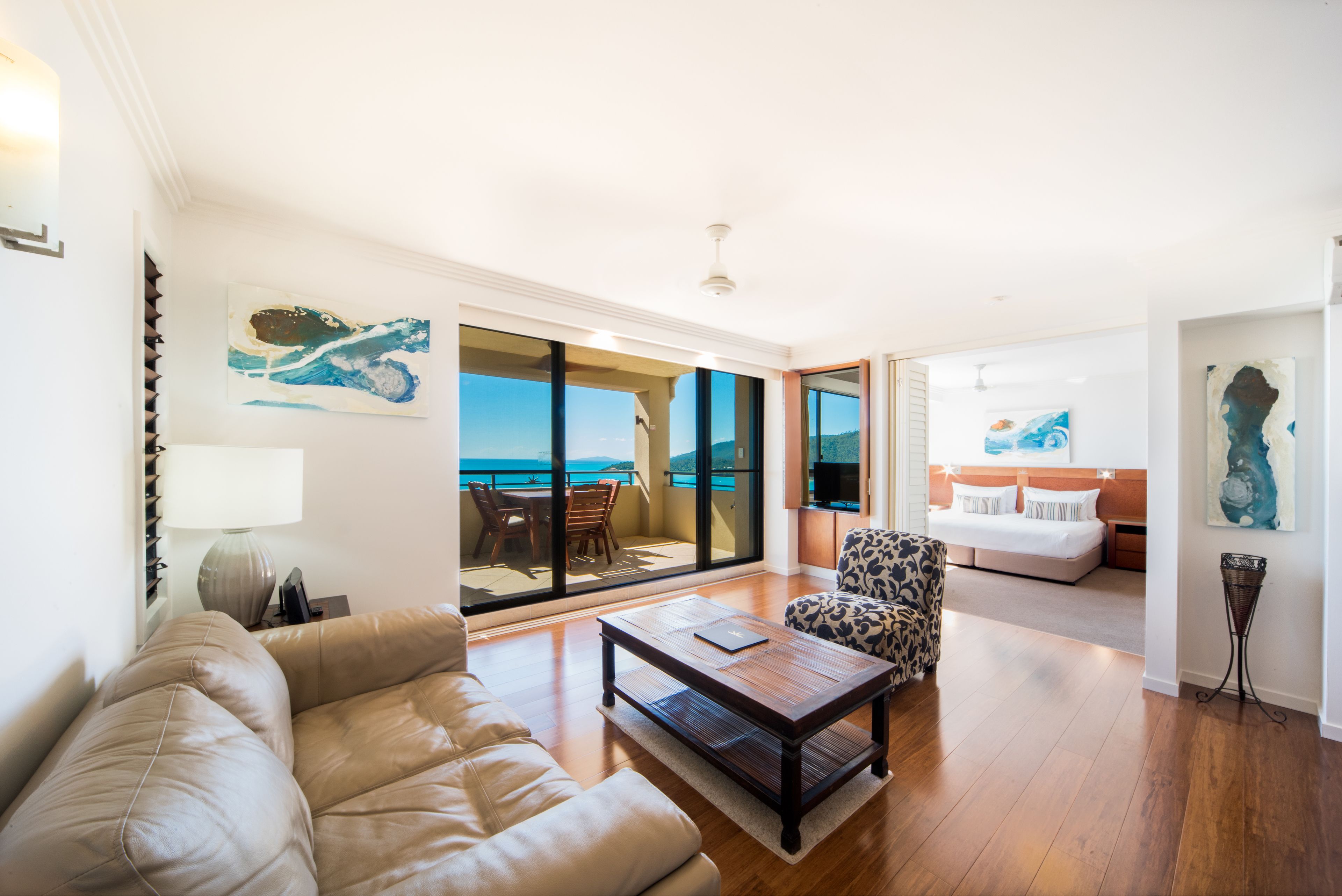 A spacious living room with a leather sofa, a patterned armchair, and a wooden coffee table. A large window offers a view of the ocean.