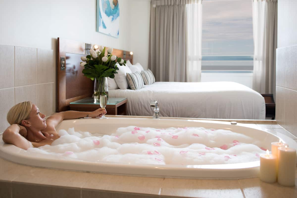 Una persona relajándose en una bañera llena de burbujas y pétalos de rosa, con una cama y una ventana con cortinas al fondo.