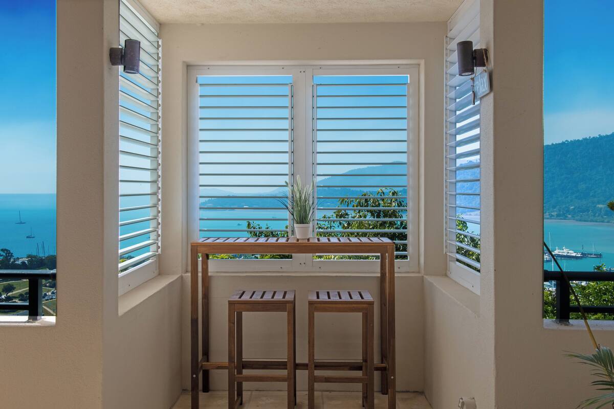 Un balcón con vistas al mar y a las montañas, con dos taburetes de madera y una planta en maceta.