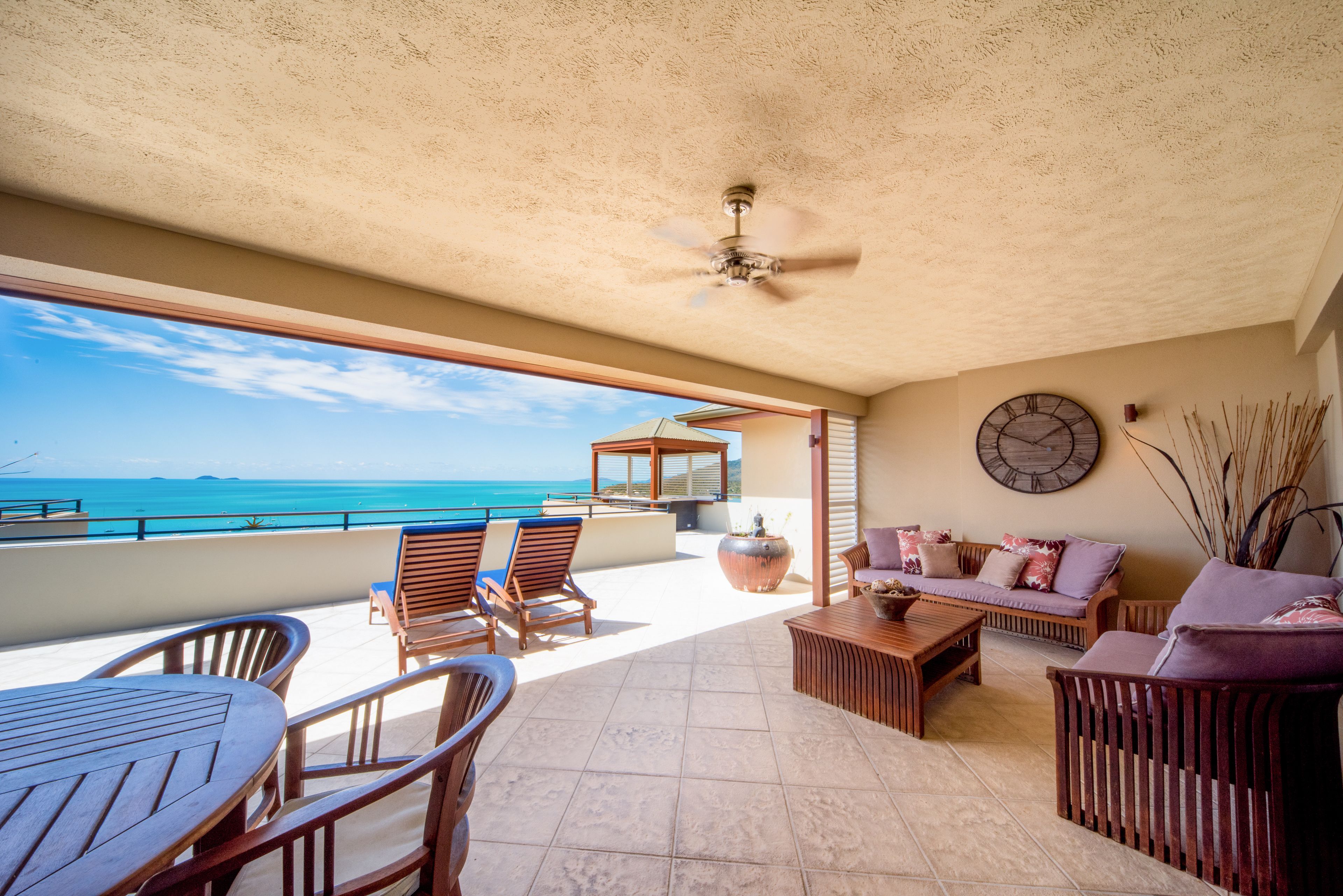 A spacious balcony with a view of the ocean, wooden furniture, and a ceiling fan.