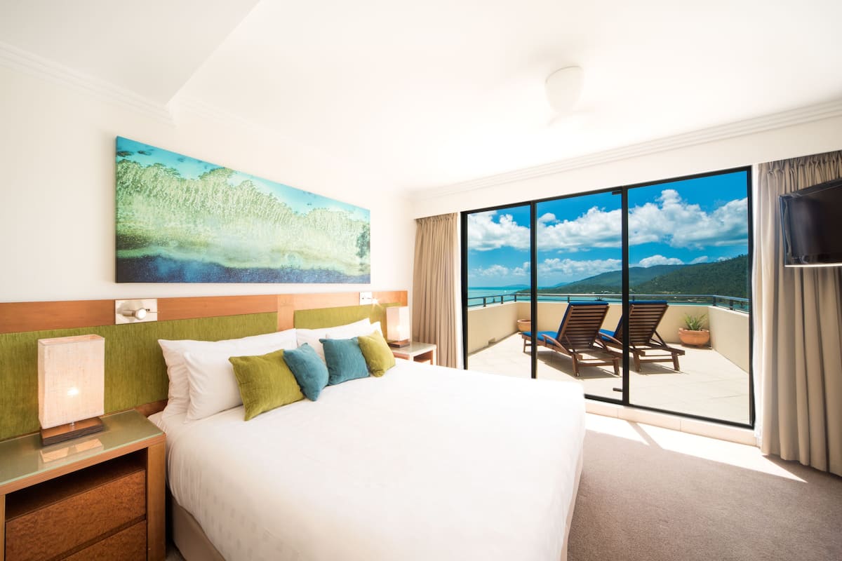 Habitación de hotel con una cama grande, un cuadro, un balcón con sillas y un televisor.