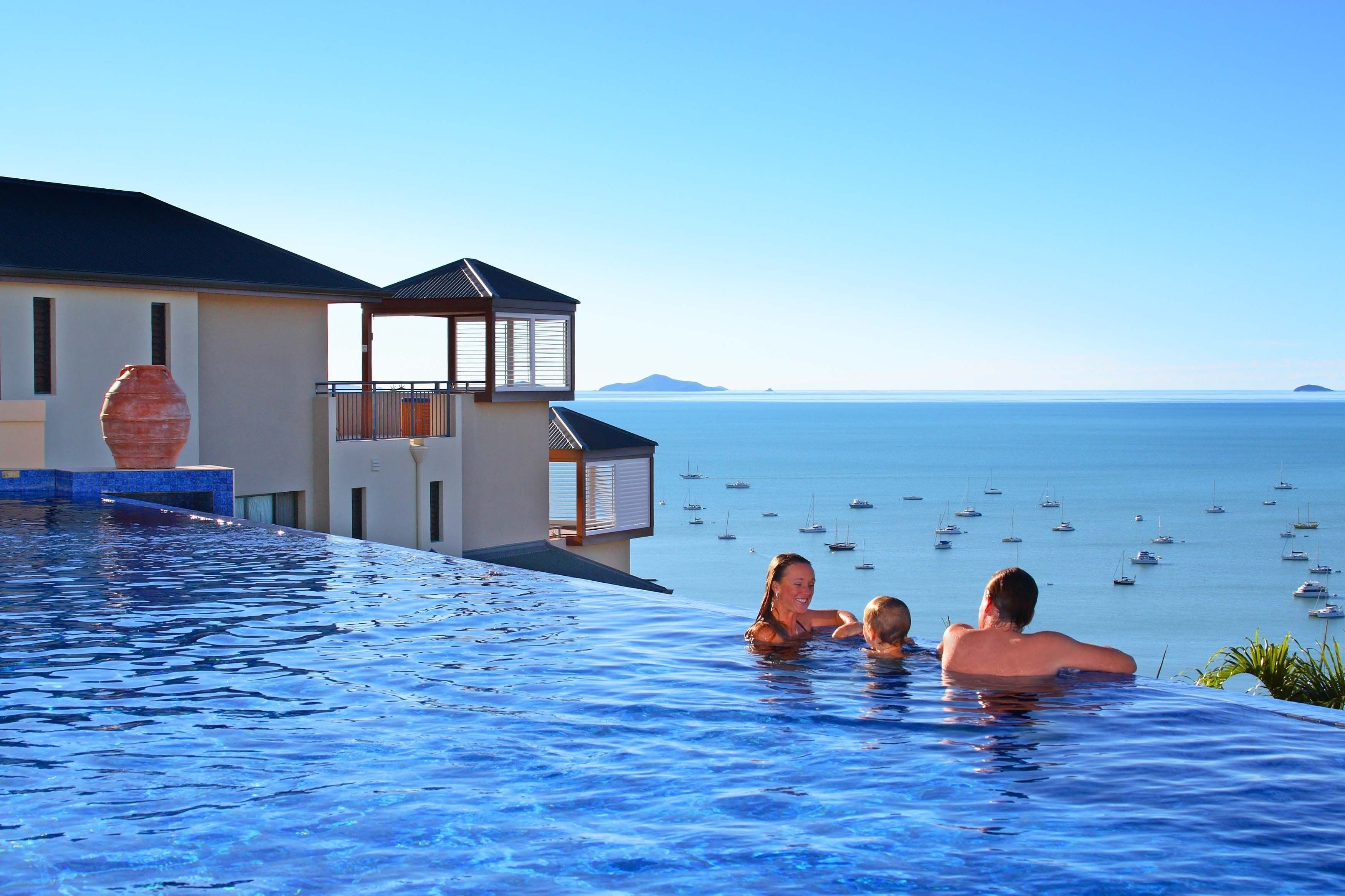 A infinity pool with a view of the sea and boats, a large vase, and a building with a balcony.