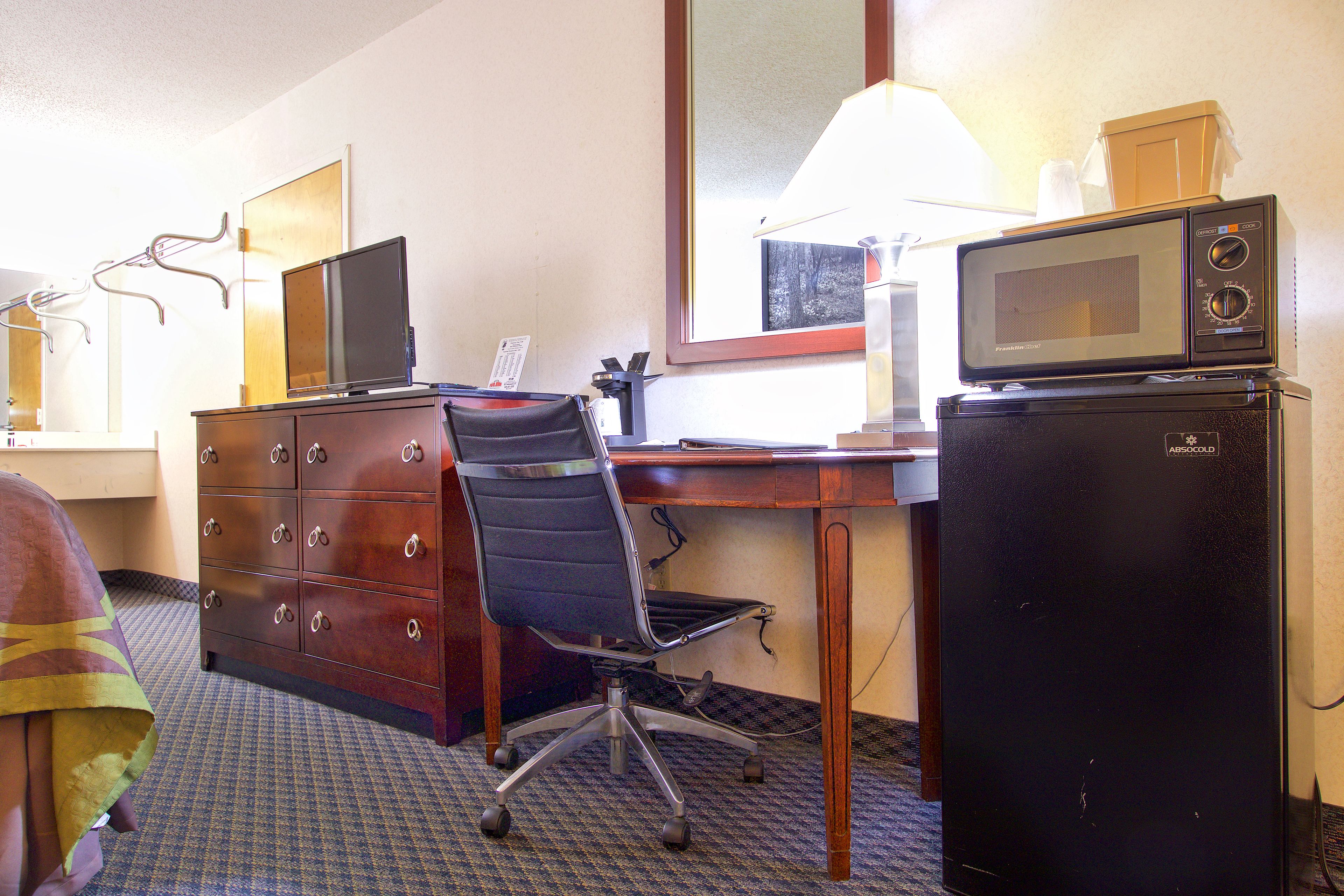 Habitación de hotel con un escritorio de madera, una silla de oficina negra, un televisor, un microondas y un refrigerador pequeño.
