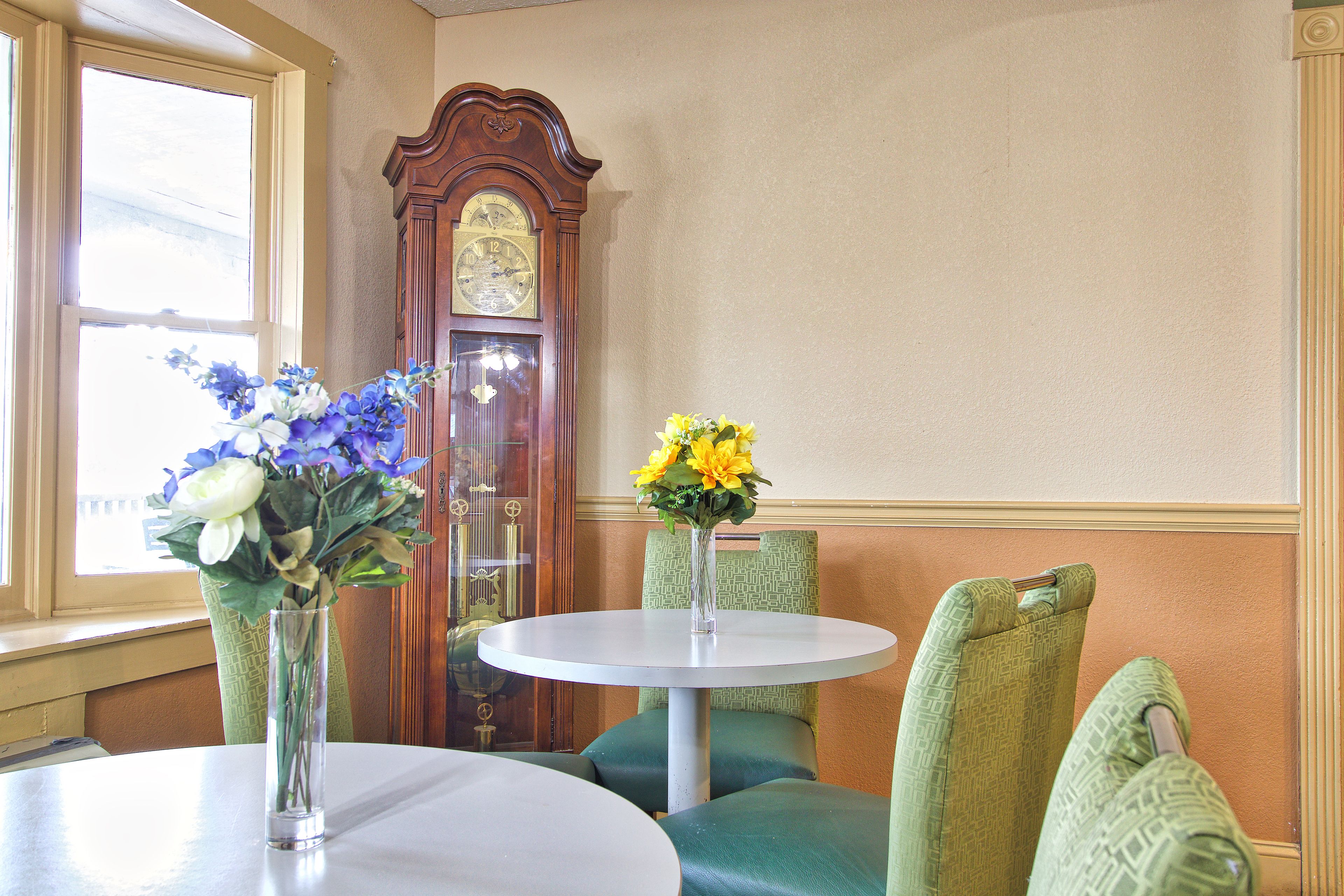 A room with a round table, two chairs, a vase with flowers, and a large wooden clock.
