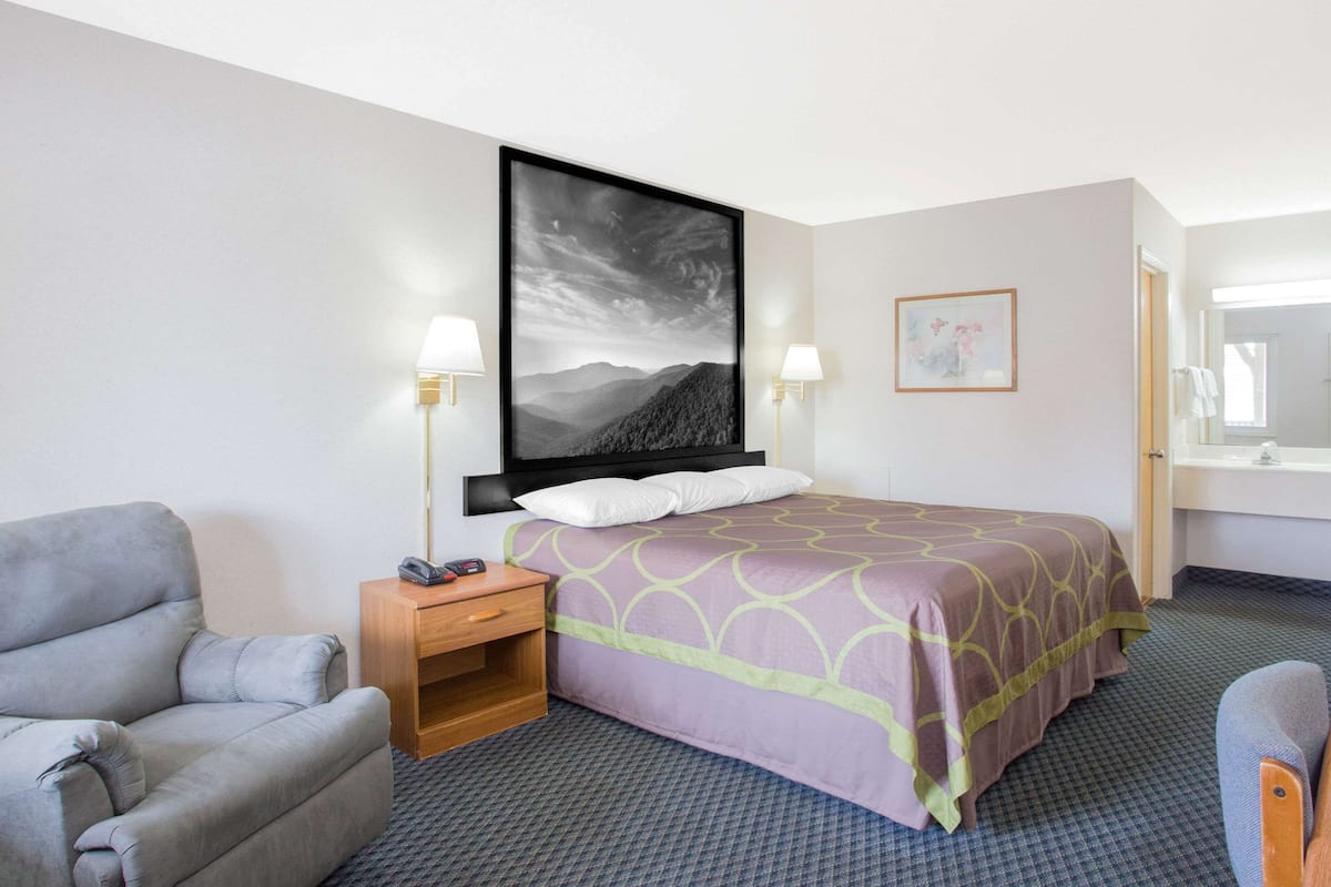 Une chambre d’hôtel avec un lit, un fauteuil gris, une table de chevet, une lampe fixée au mur et un tableau encadré.