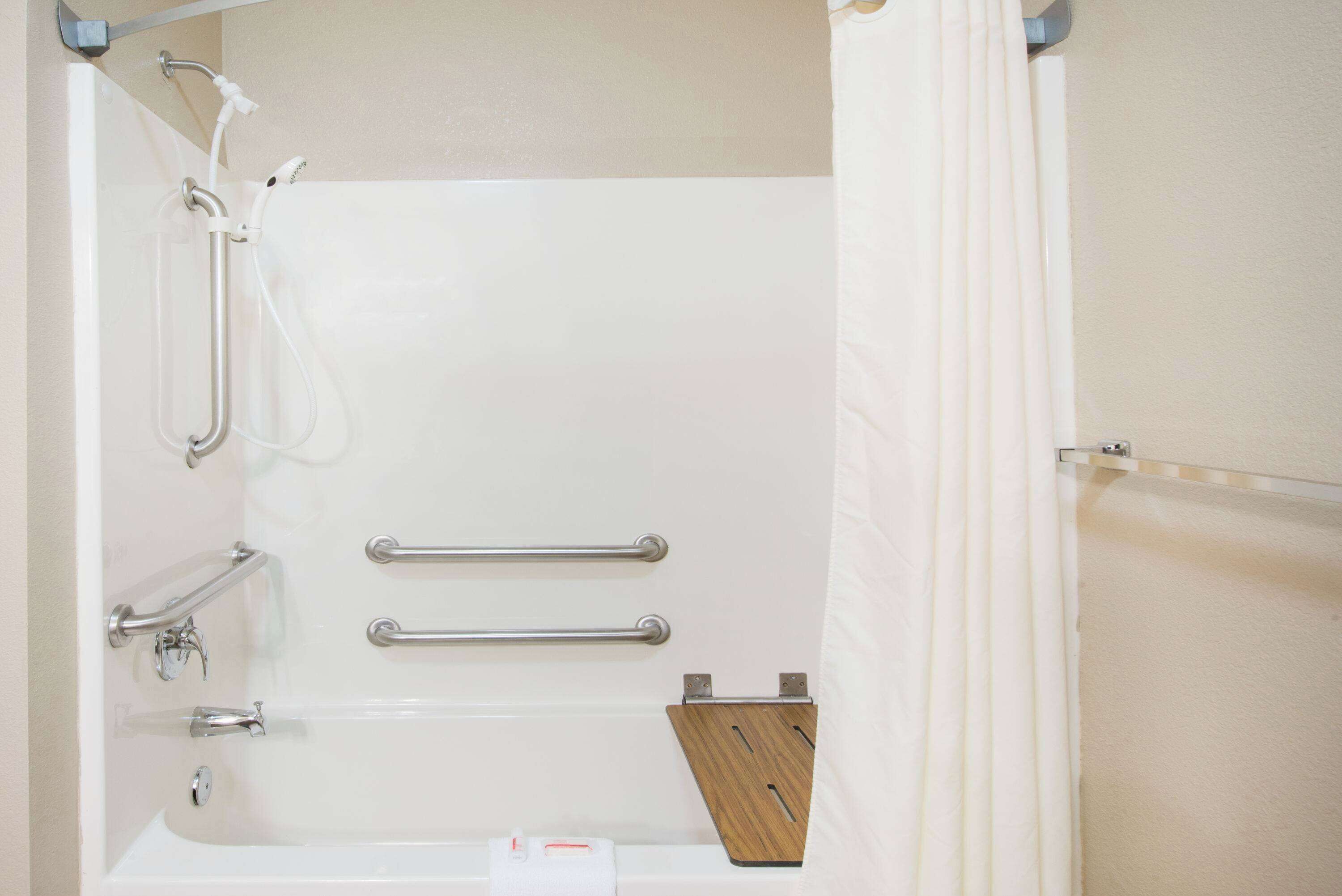 A bathtub with grab bars, a shower hose, and a wooden shelf.