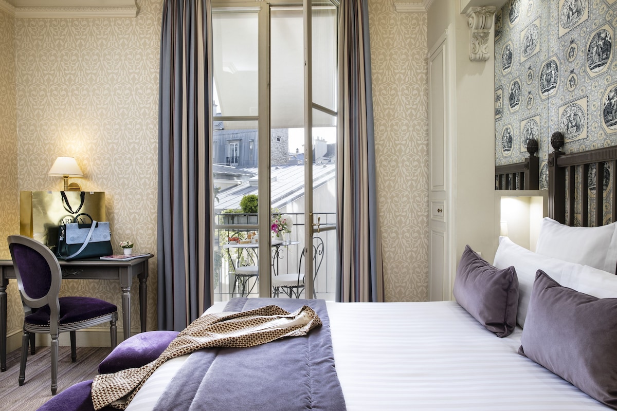 Habitación de hotel con cama, escritorio con lámpara, silla y vista de la ciudad a través de un ventanal grande.