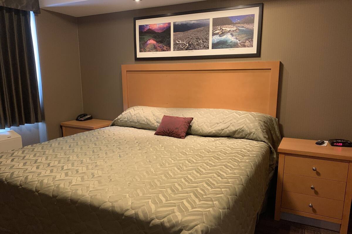 A hotel room with a large bed, a nightstand with a phone, a digital clock, and a framed picture above the bed.