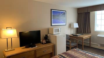 Habitación de hotel con cama, escritorio, televisor, microondas, lámpara, silla y ventana con cortinas.