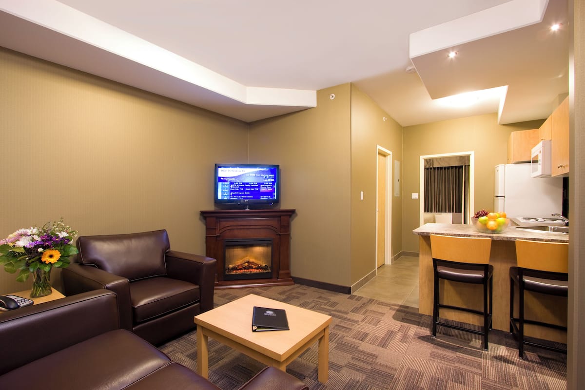 A modern hotel room with a living area, a fireplace, a television, a small kitchen area with a fruit bowl, and a dining table with two chairs.